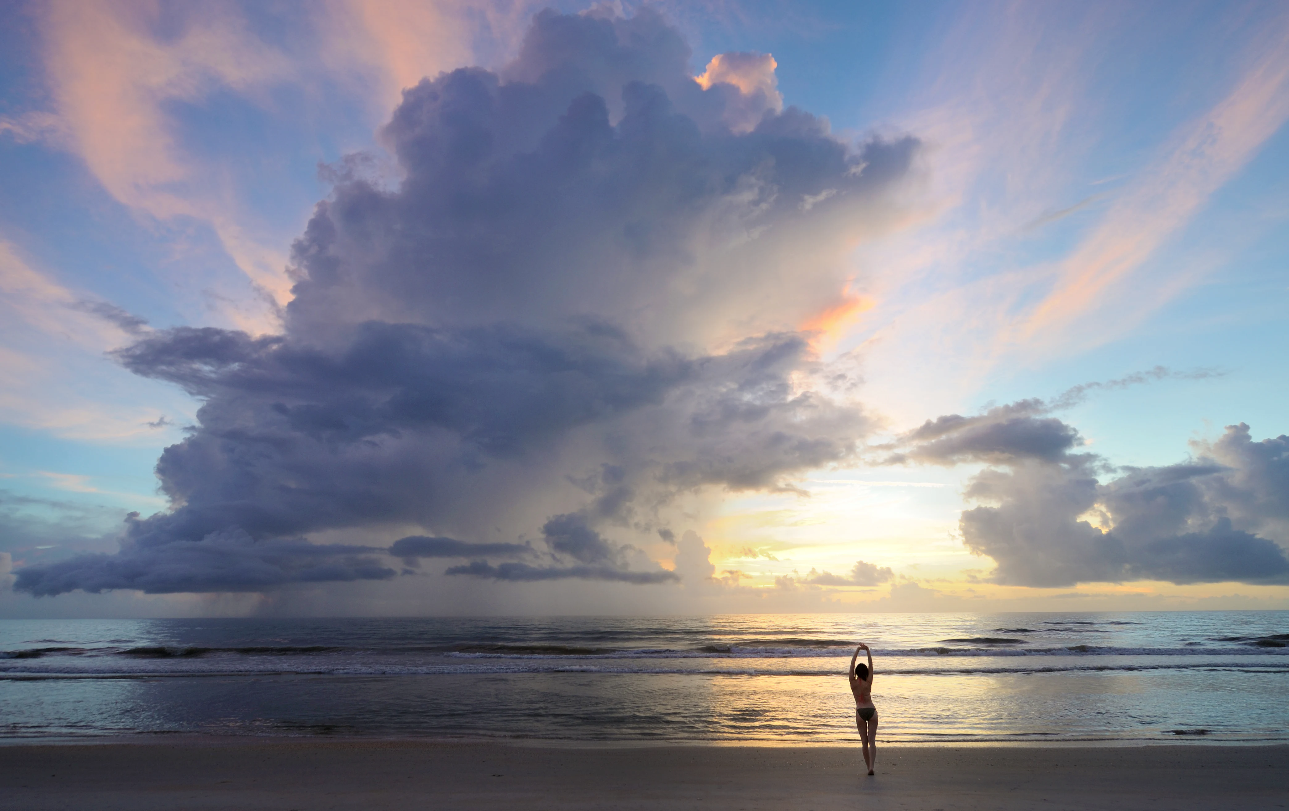 A wide shot of a person standing with arms raised on a sandy beach at sunrise, with a large, dark, rain-filled cloud formation over the ocean.
