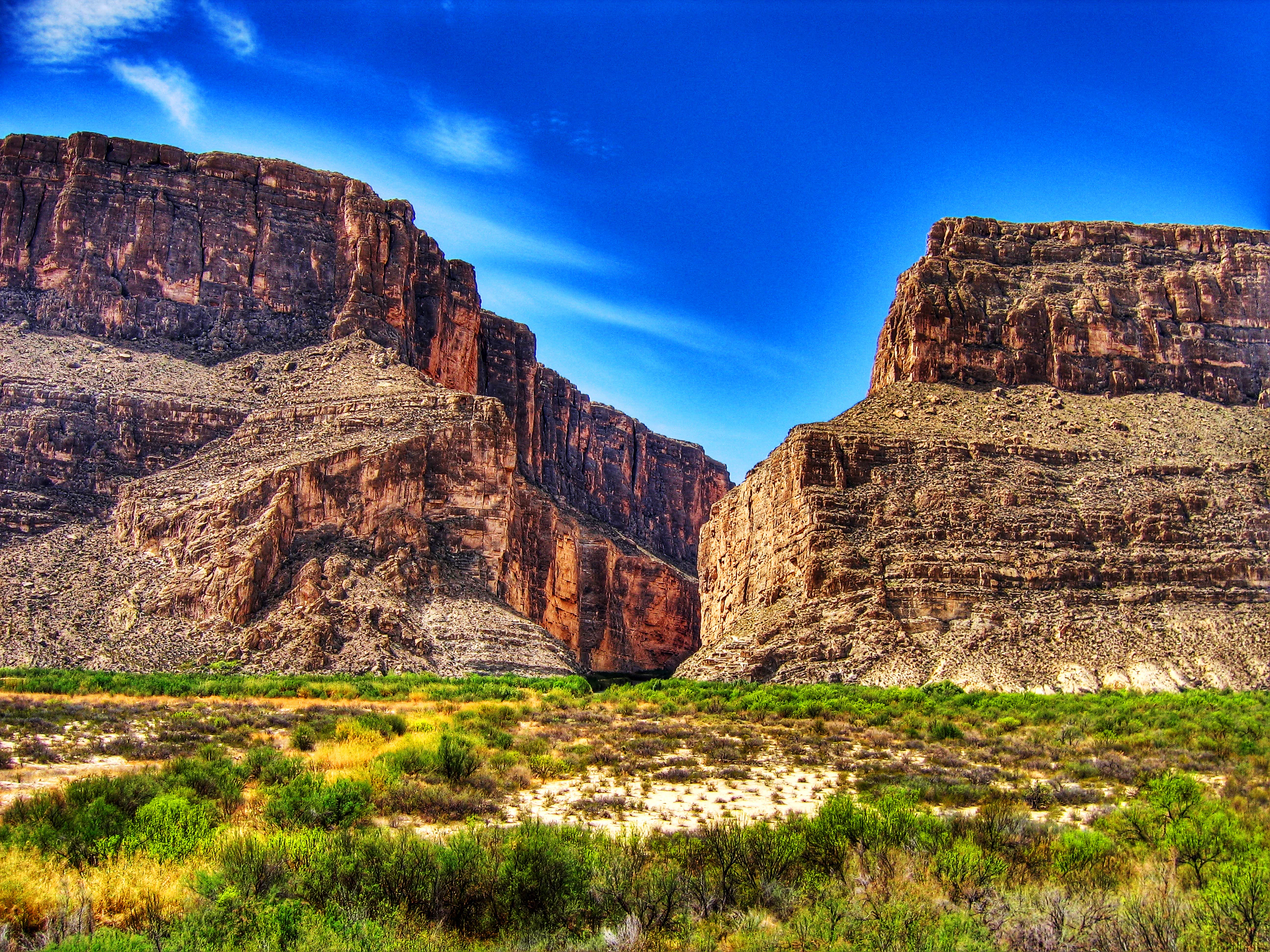 A wide, low-angle shot of a deep canyon with massive, layered limestone rock walls rising on either side, separated by a narrow gap, with arid desert vegetation and shrubs in the foreground under a vibrant blue sky.