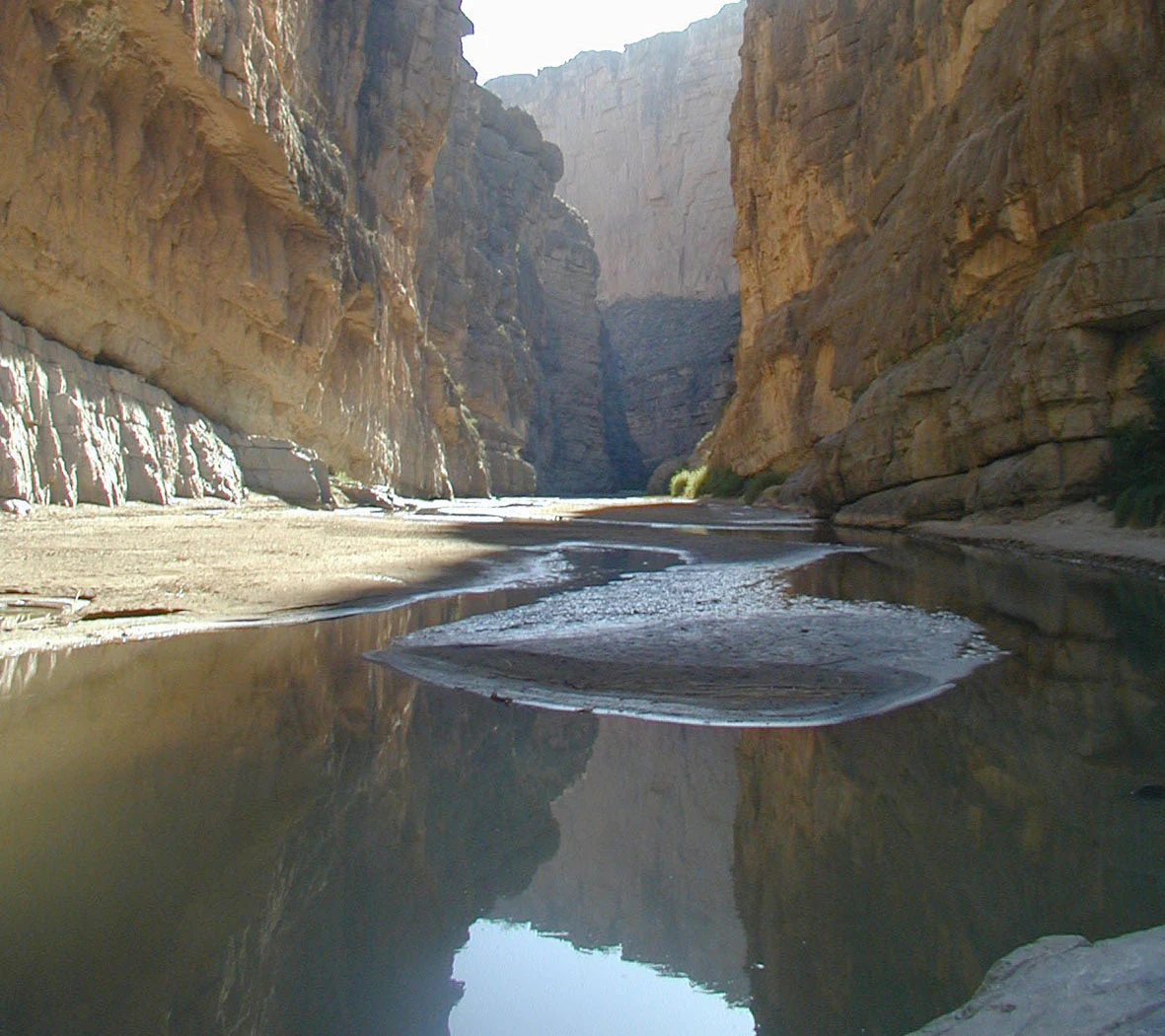 A view of a river winding through a deep limestone canyon with steep, high rock walls reflecting in the calm water.