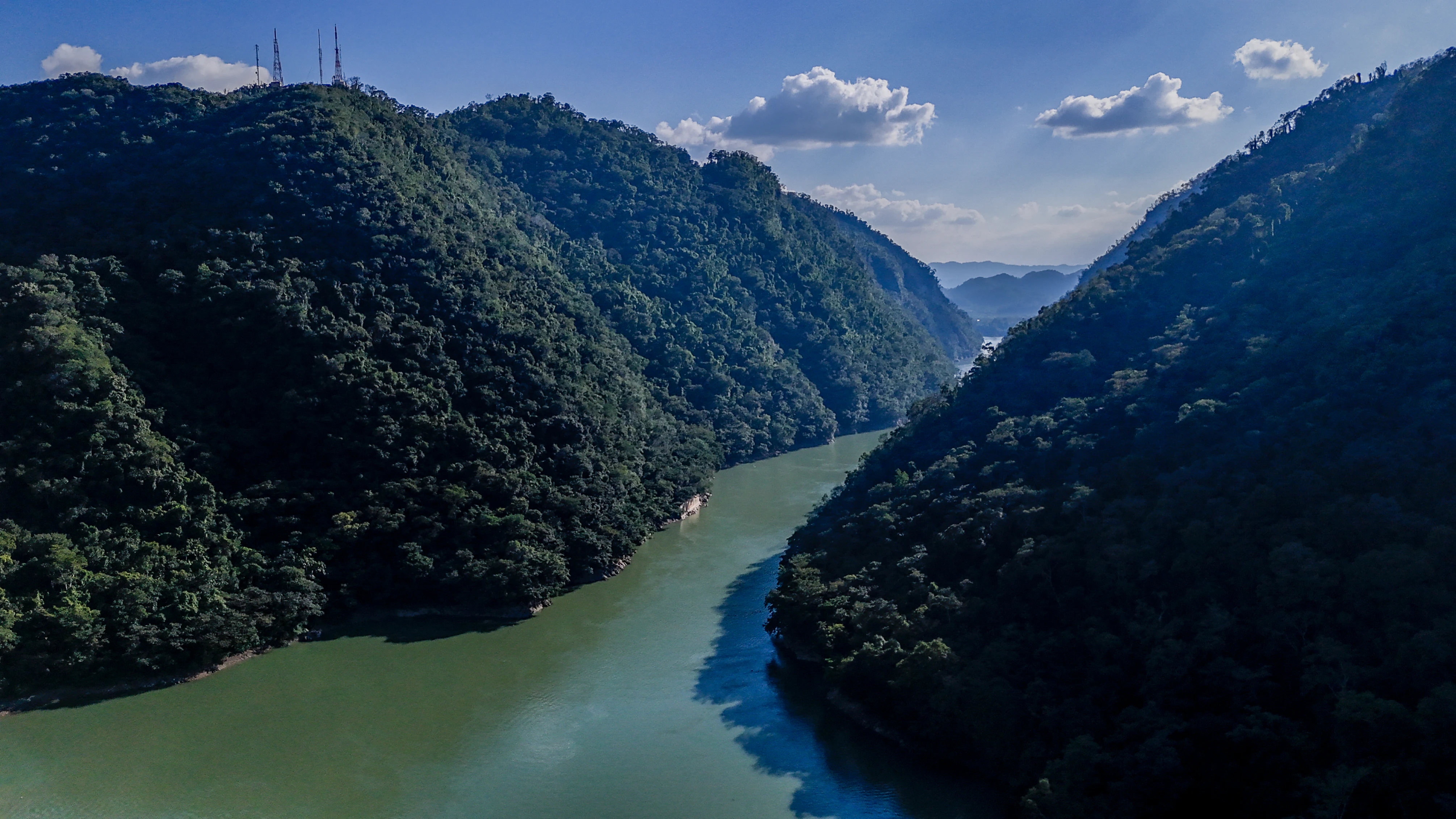 Aerial drone perspective looking down a winding river between two steep, heavily forested canyon walls under a clear blue sky.