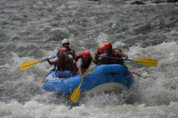 A blue inflatable raft carrying five people wearing helmets and life jackets navigates through turbulent river rapids.