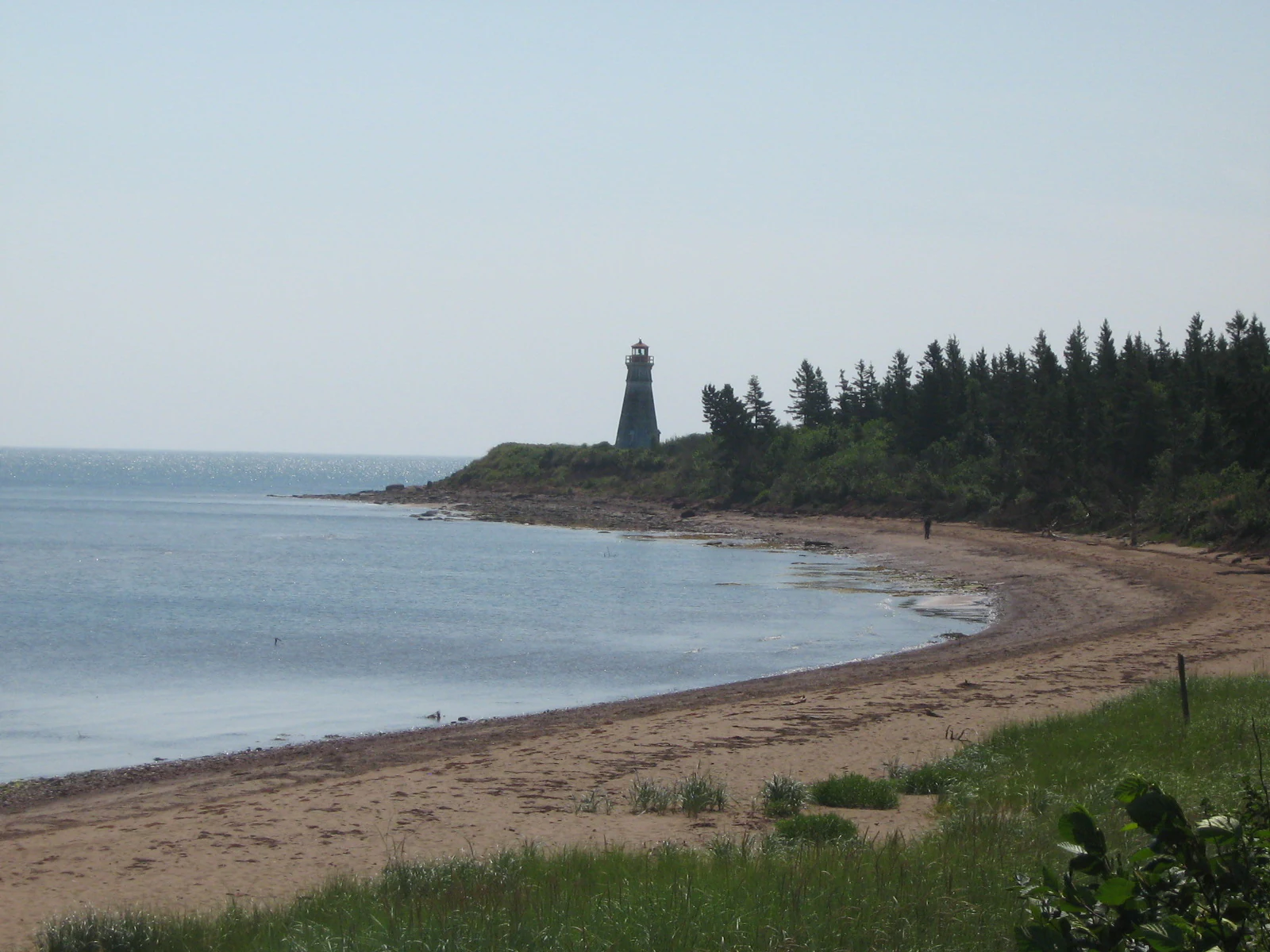 A distant view of the Cap Jourimain lighthouse perched on a grassy point above a sandy beach and calm coastal water.