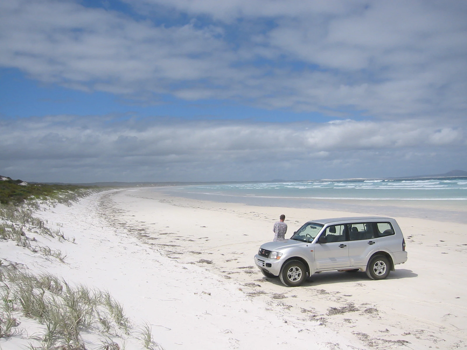 SUV vehicle parked on white sandy beach with grassy dunes, ocean waves, and a person standing near the vehicle under a partly cloudy sky