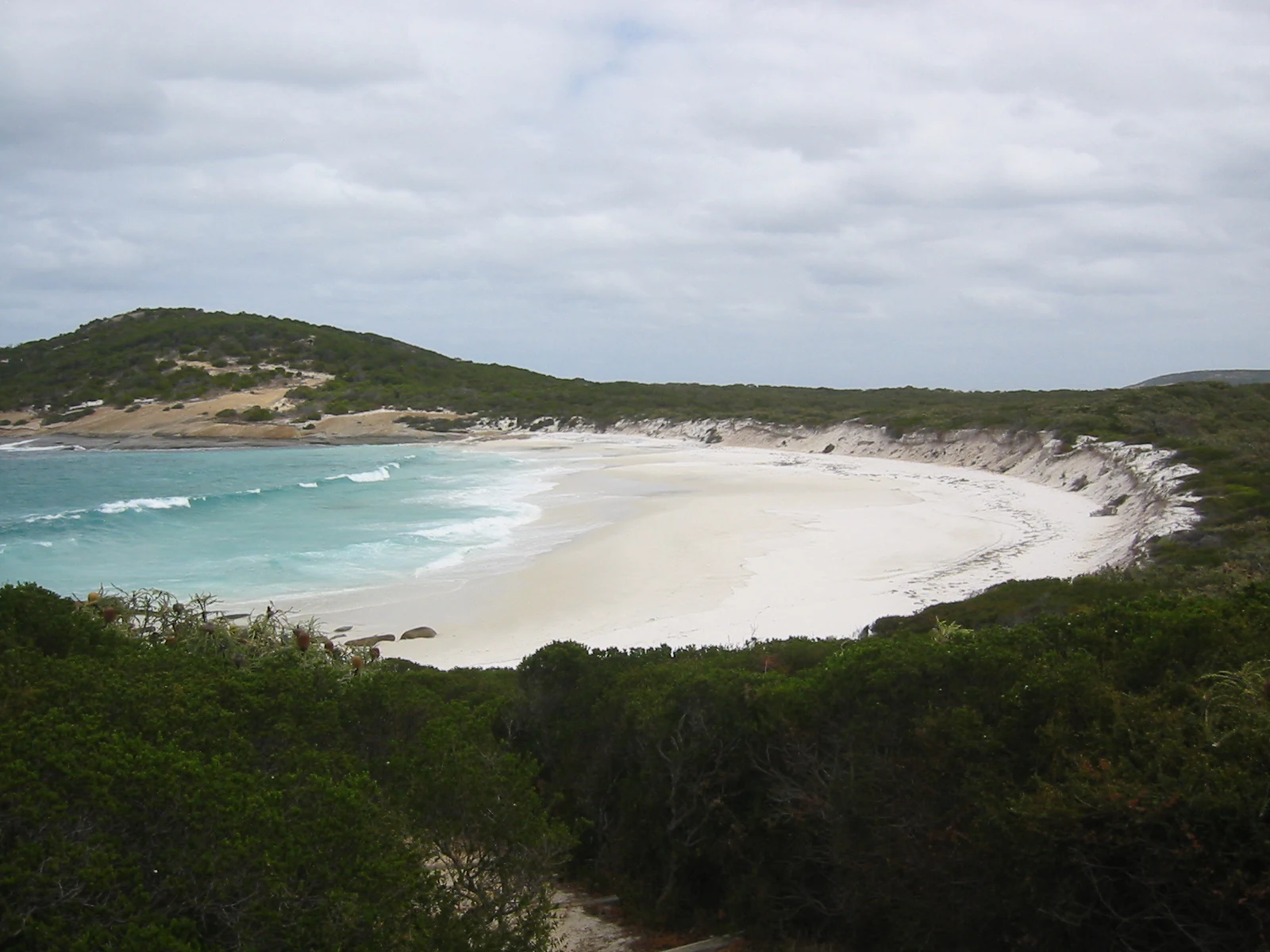 A coastal bay with turquoise water, sandy beach, and green vegetation under a cloudy sky