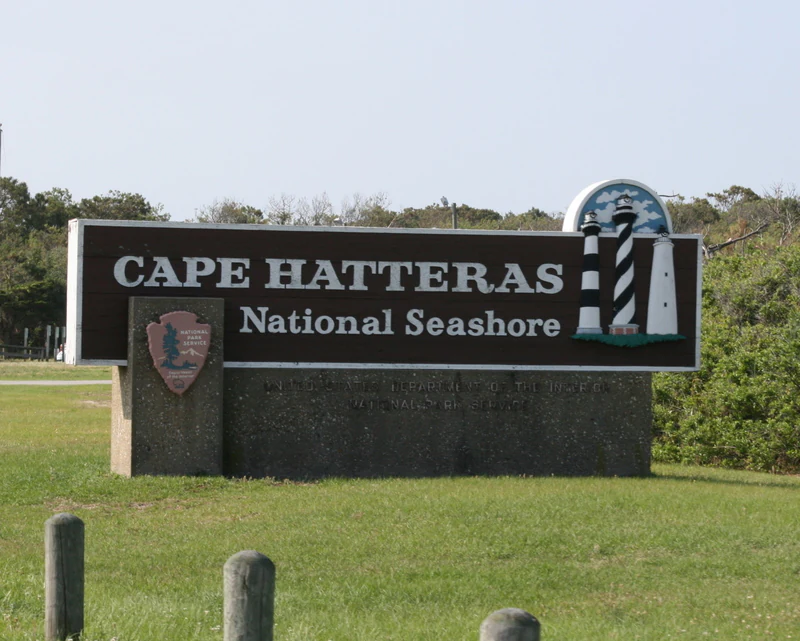 A brown rectangular park entrance sign for Cape Hatteras National Seashore featuring the National Park Service arrowhead logo and an icon of a striped lighthouse.