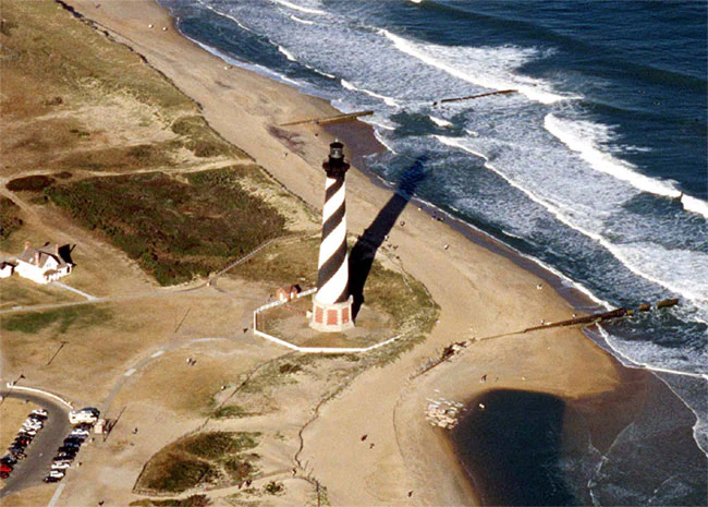 An aerial shot of the Cape Hatteras Lighthouse standing on a sandy beach beside the Atlantic Ocean, with a parking area visible on the lower left.