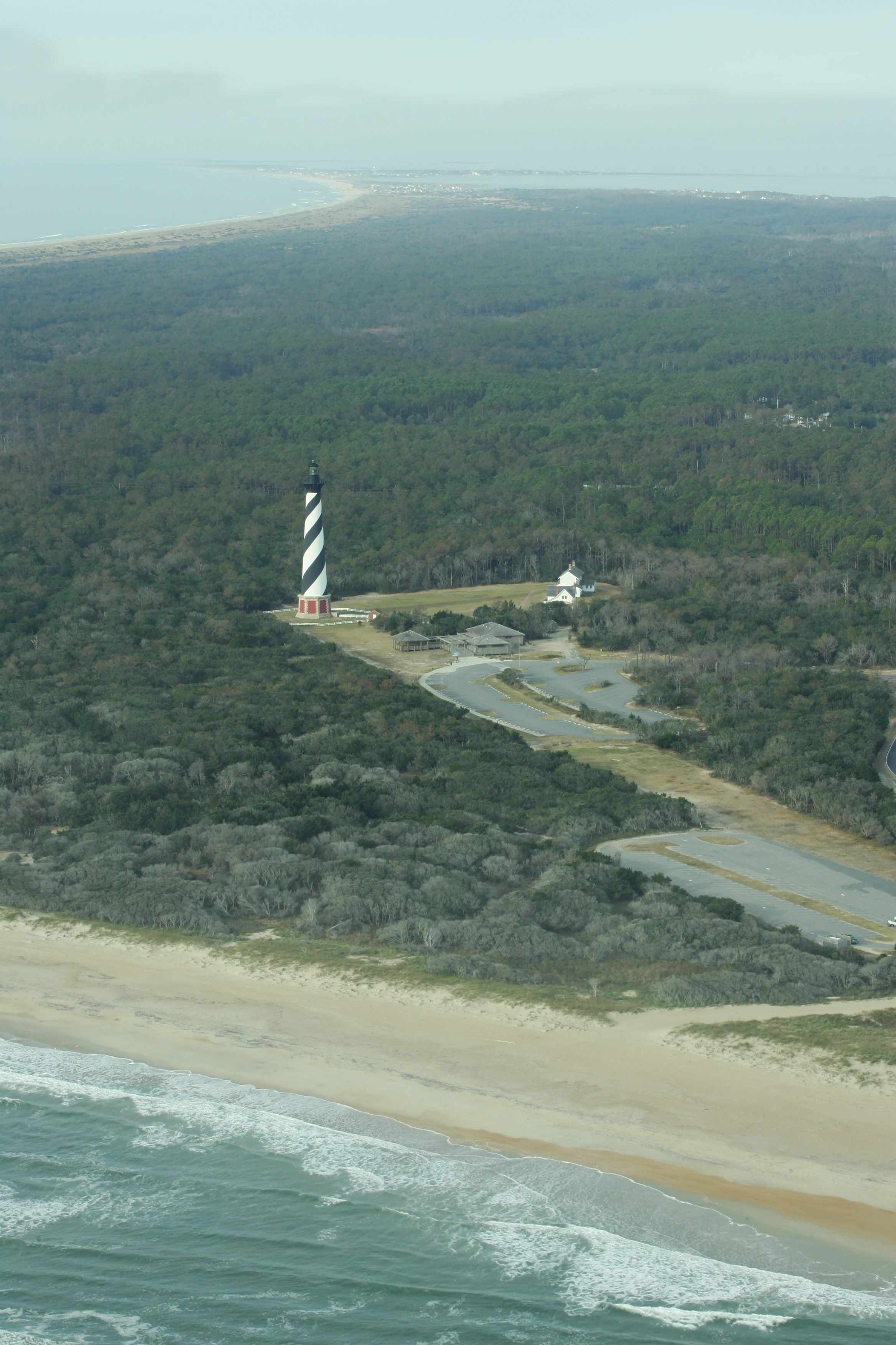 An aerial photograph showing the striped Cape Hatteras Lighthouse situated amidst dense maritime forest with a sandy coastline and the Atlantic Ocean in the foreground.