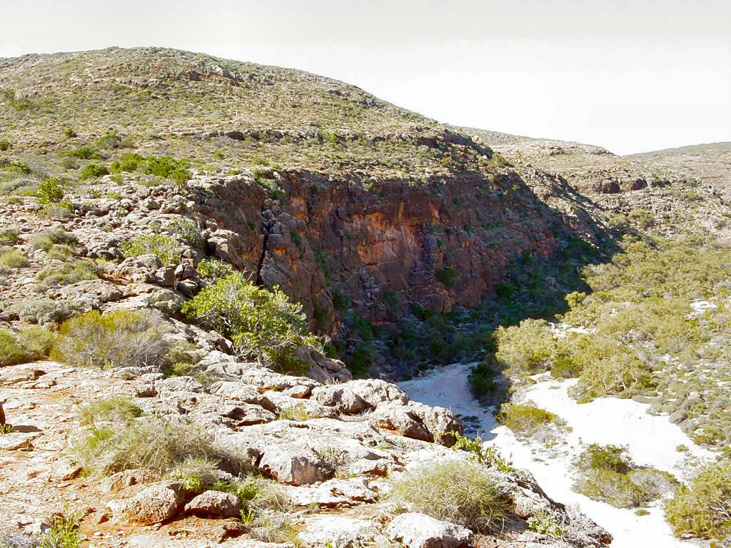 Rocky canyon landscape featuring steep cliffs, scattered shrubs, and a narrow waterway flowing through rugged terrain