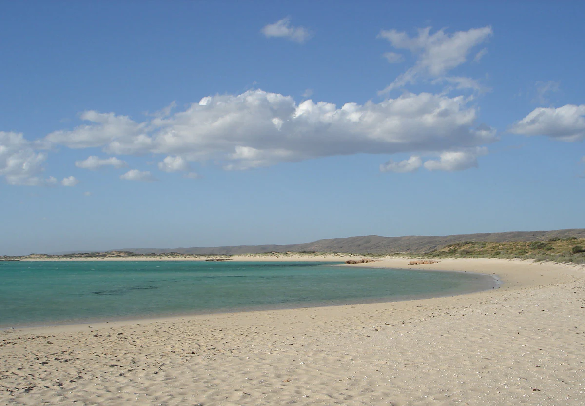 Sandy beach with turquoise water, scattered clouds, and distant hills under a clear blue sky.