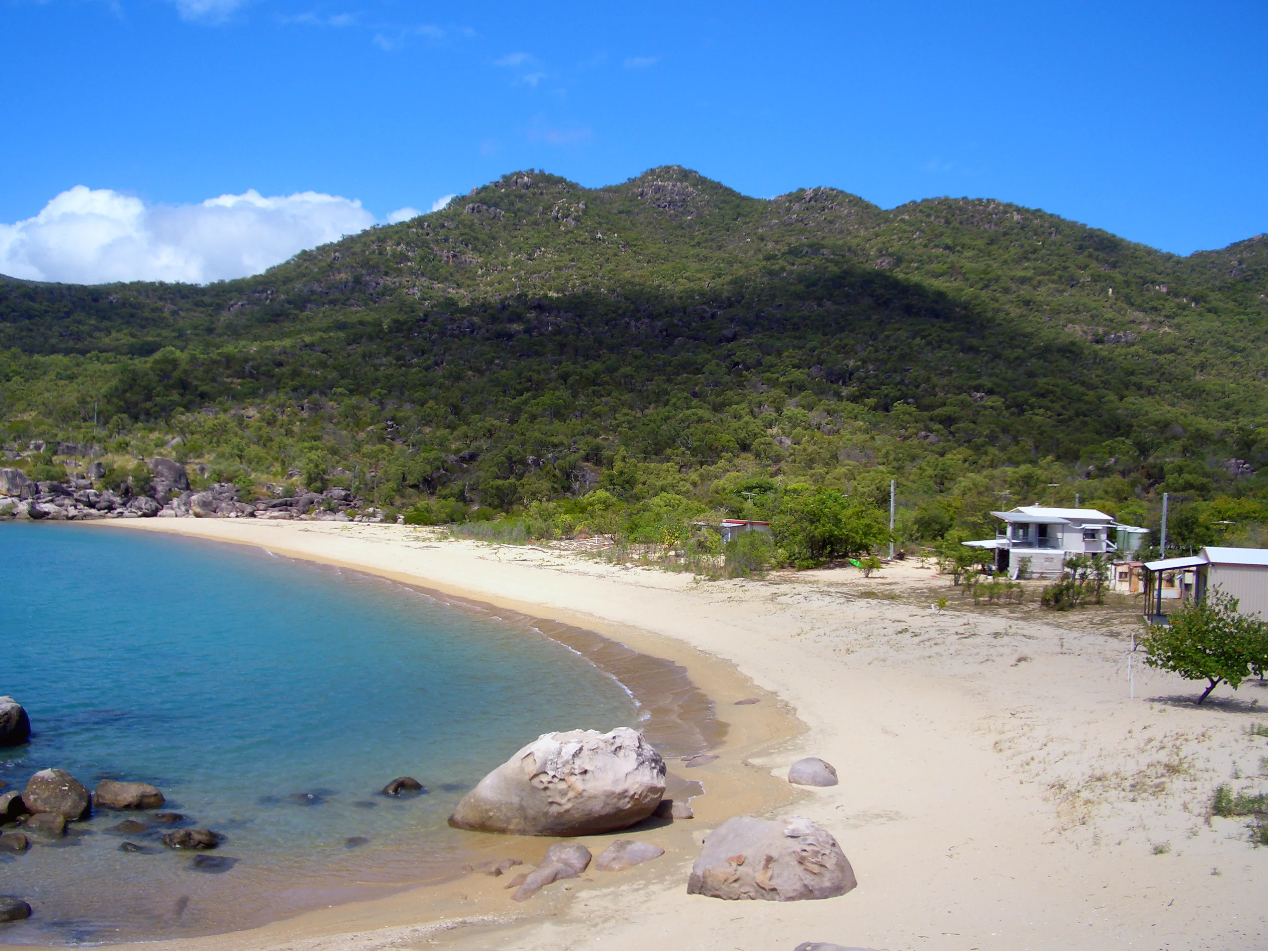 Playa arenosa con aguas turquesas, grandes rocas, terreno montañoso verde y algunos edificios bajo un cielo azul despejado
