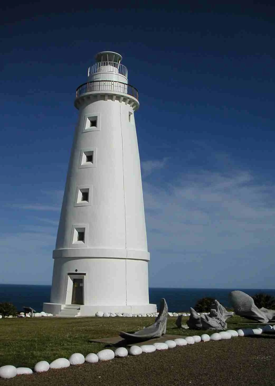 A white tapered lighthouse stands against a clear deep blue sky, with the ocean visible in the background and a stone-lined garden path in the foreground.