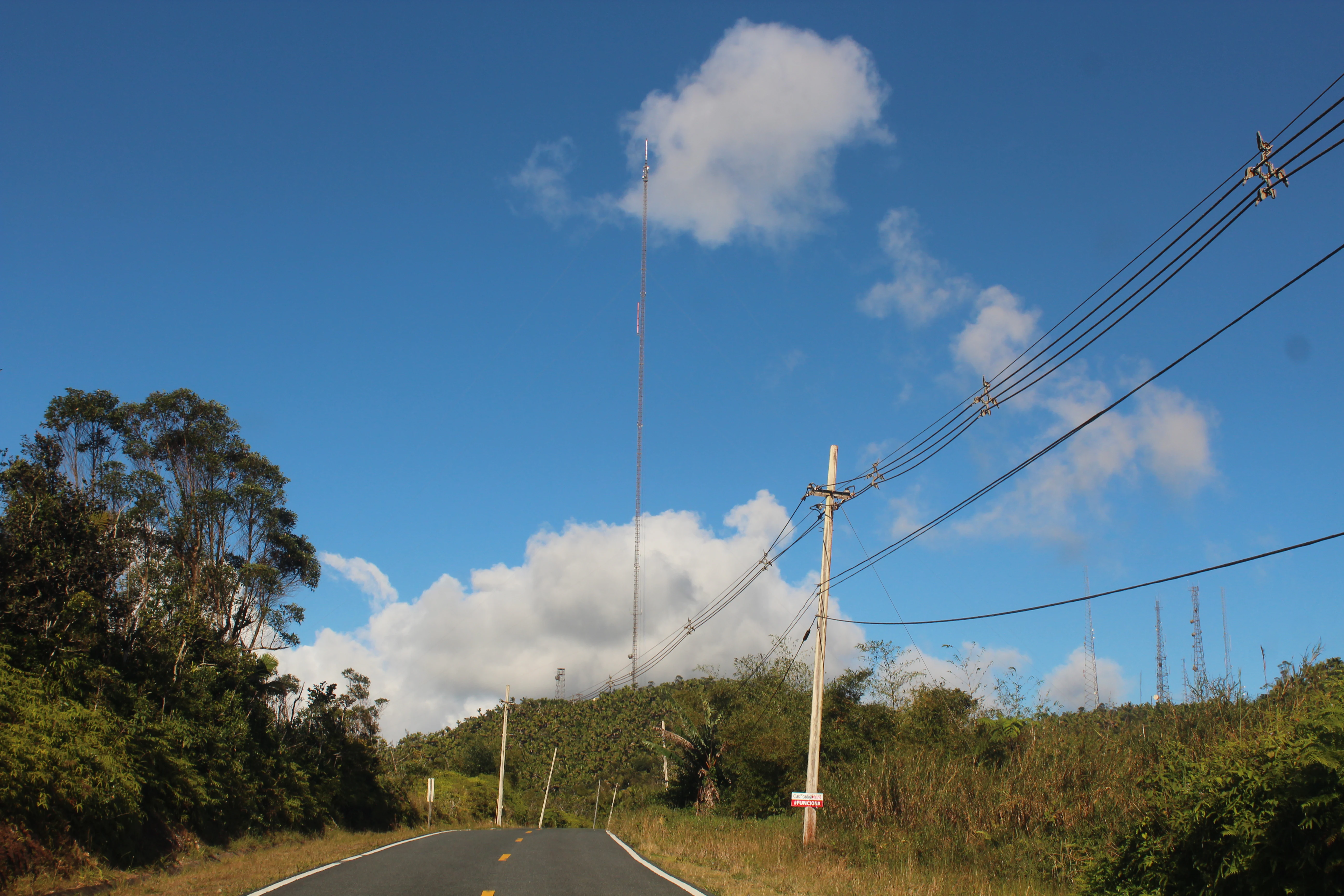 A paved mountain road winds forward, flanked by dense green forest on the left and scrubby vegetation on the right, with tall communication towers visible against a blue sky.