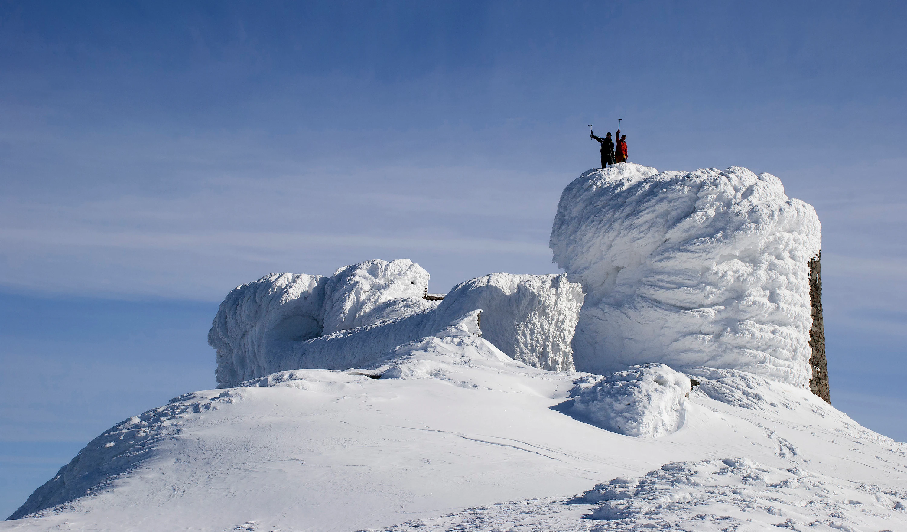 Snow-covered rocky mountain summit with two people standing on top against clear blue sky