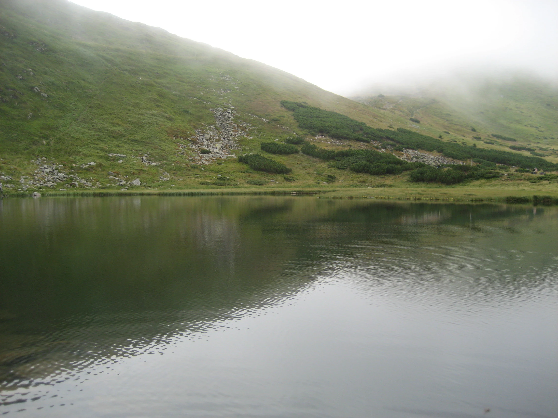 Calm lake reflecting green mountain slopes under overcast skies with low-lying clouds