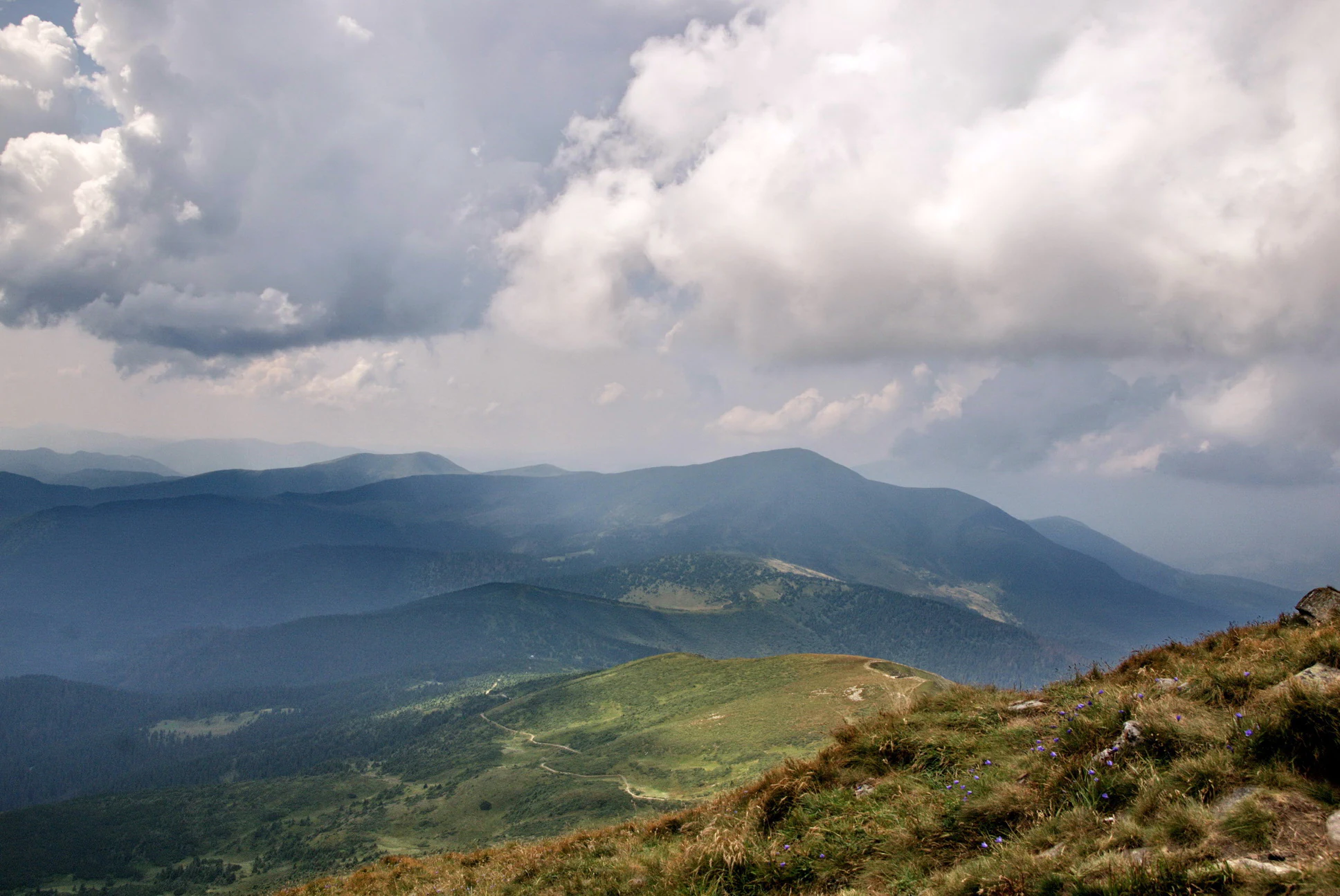 Grassy hillside with rolling mountain ranges extending into the distance under a partly cloudy sky