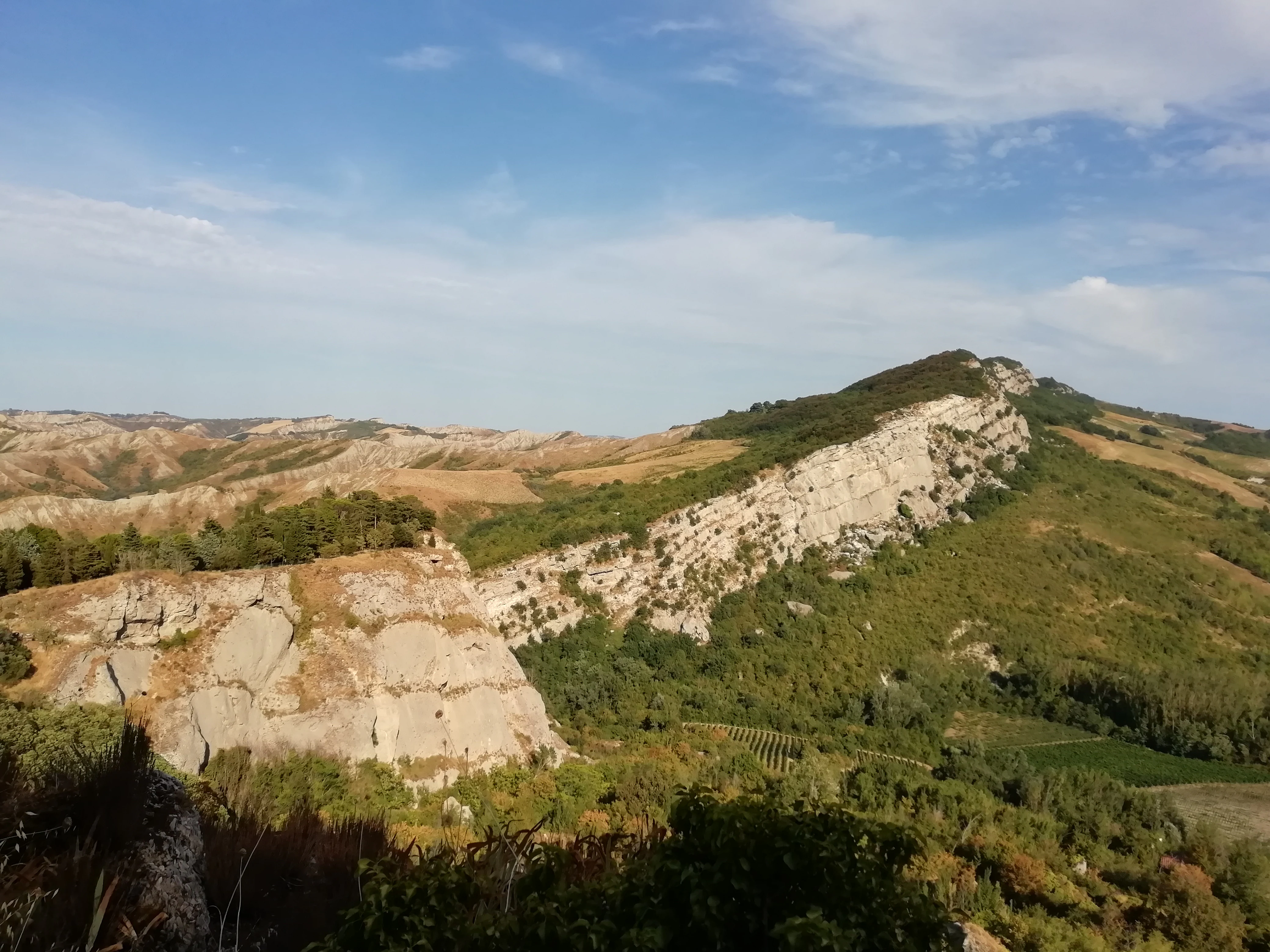 A landscape view of light-colored, exposed gypsum rock formations and rugged hills in the Italian Appennine region under a clear sky.