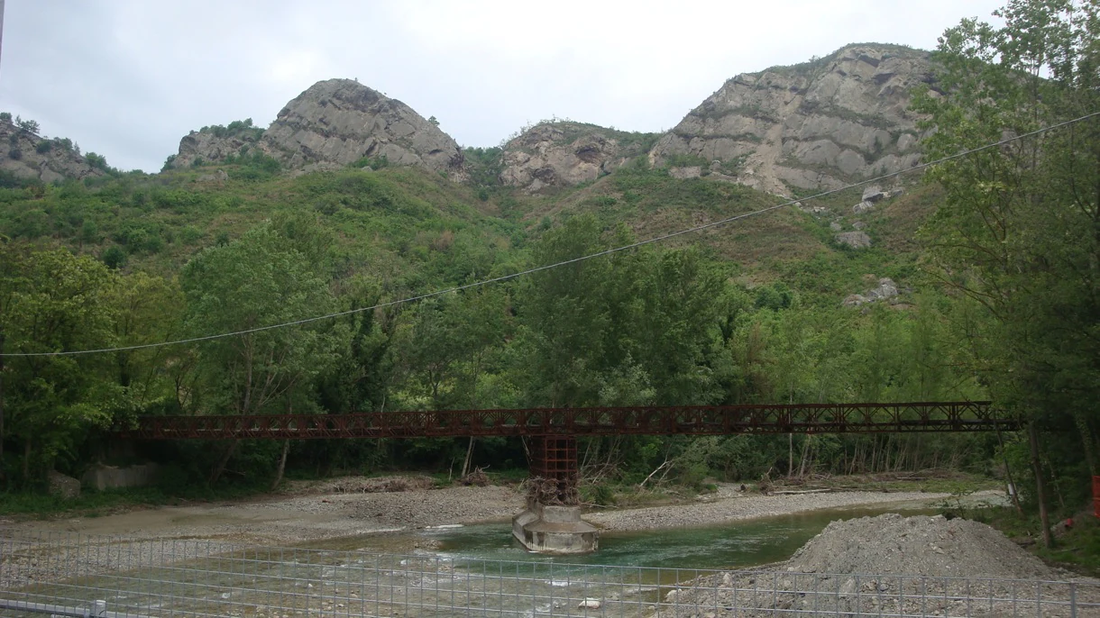 A metal truss bridge crossing a shallow, rocky river with green hills and exposed geological cliffs in the background.