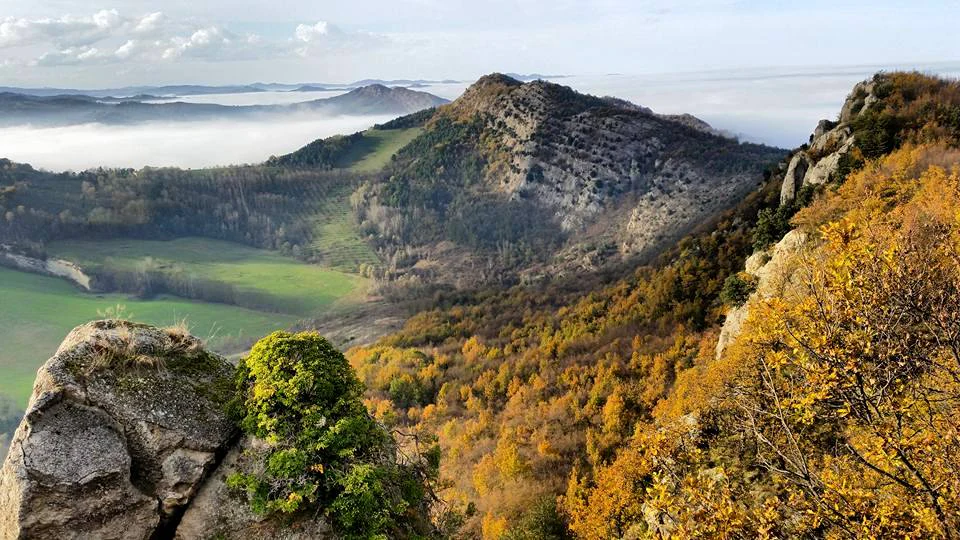 A wide angle landscape shot of the Vena del Gesso Romagnola ridge in autumn, showing rocky outcrops, vibrant orange and yellow forest foliage, and a valley filled with low mist.