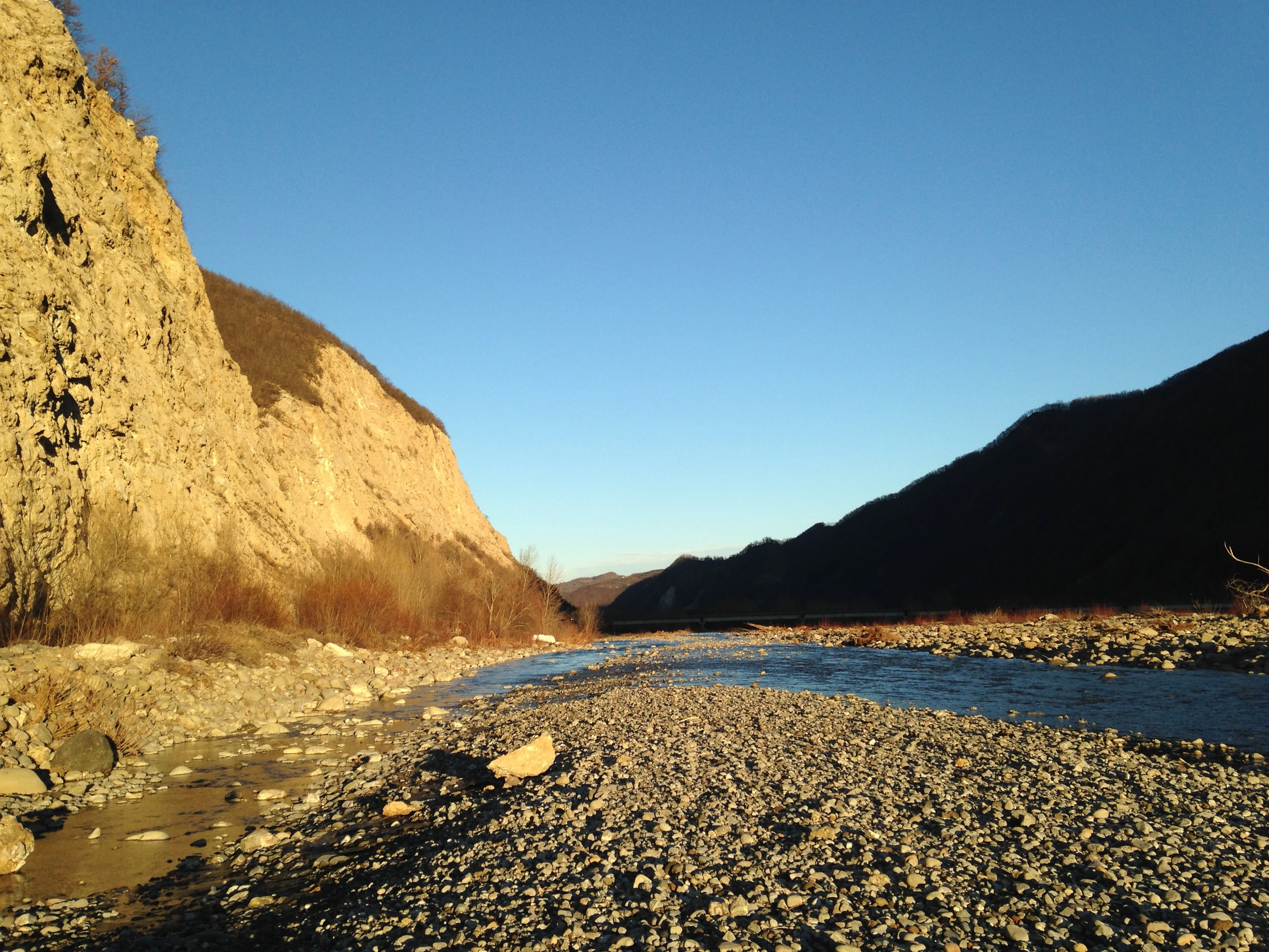 A wide angle view of the Secchia River flowing between steep, sunlit gypsum cliffs and rocky embankments under a clear blue sky.