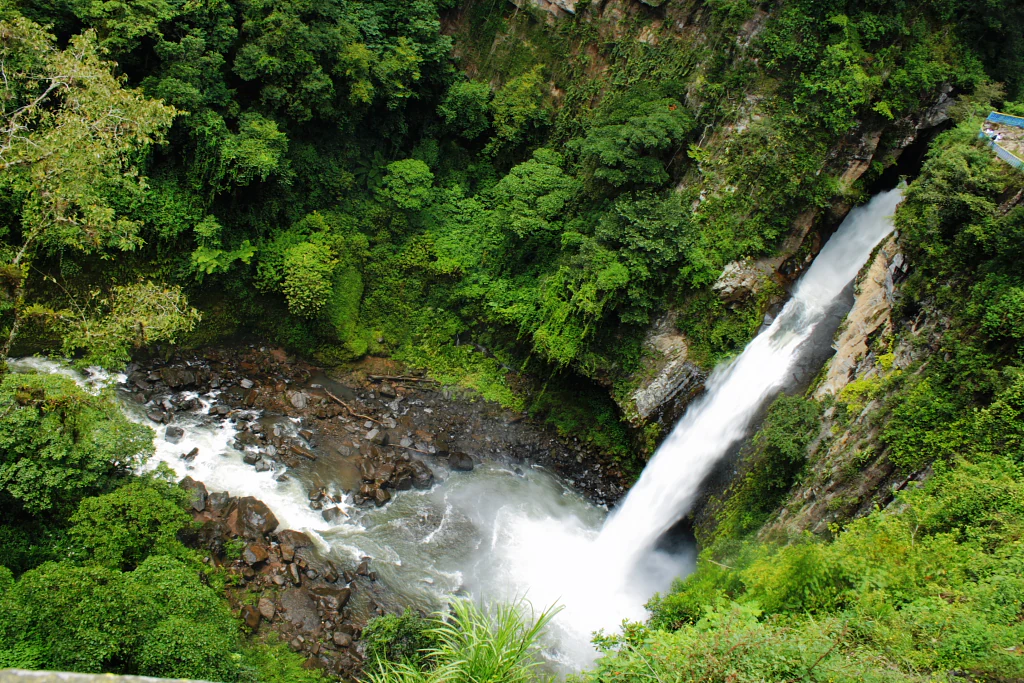 A high-angle view of a large waterfall plunging into a pool within a dense, green forest canyon.