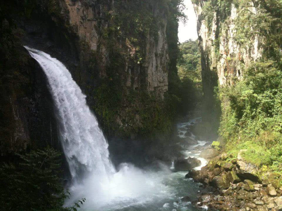 A large waterfall flows down a rocky cliff into a river running through a steep, lush, forested gorge.