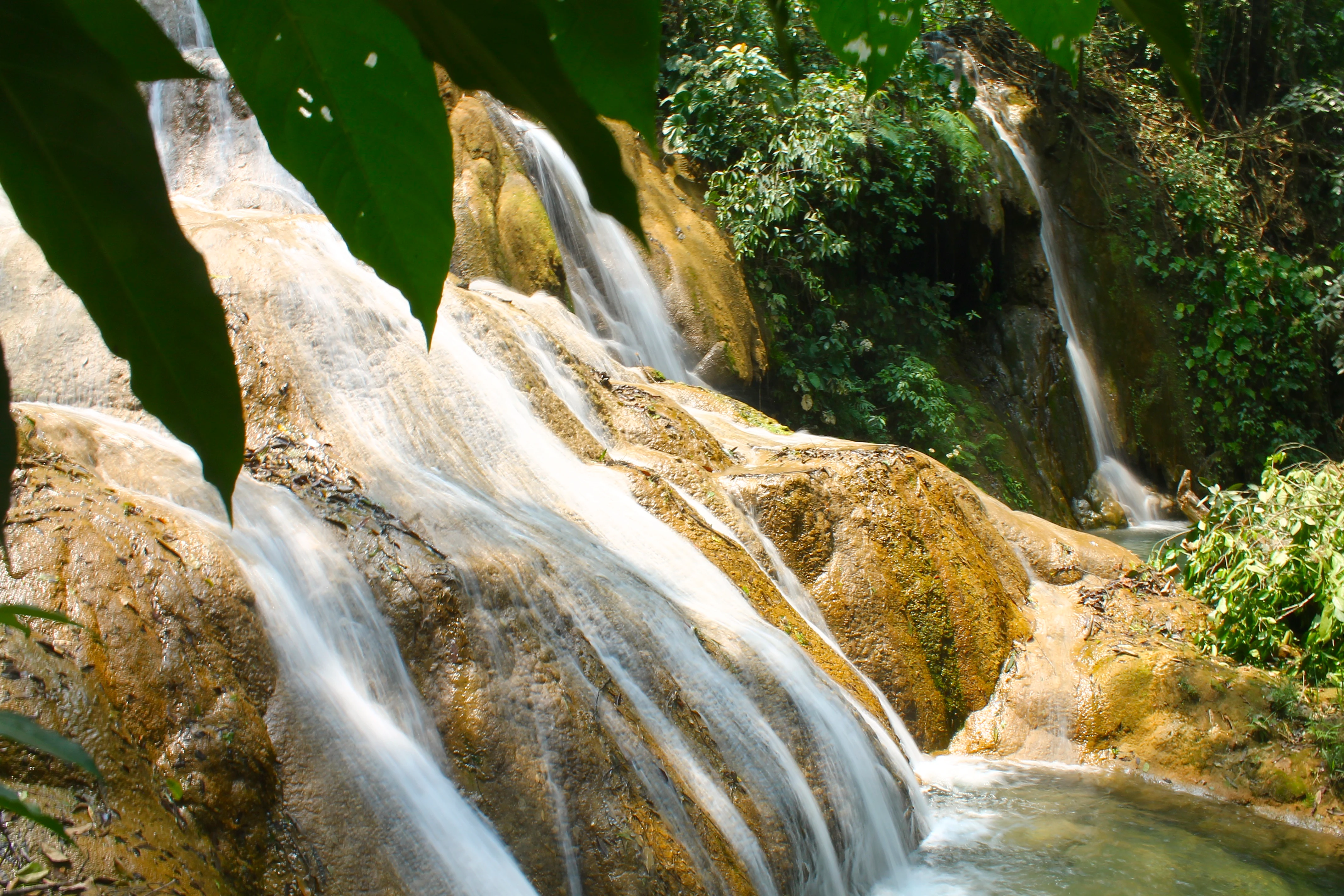 A close-up view of tiered limestone waterfalls cascading over light-brown rocks into a pool, framed by lush green jungle foliage in the foreground.