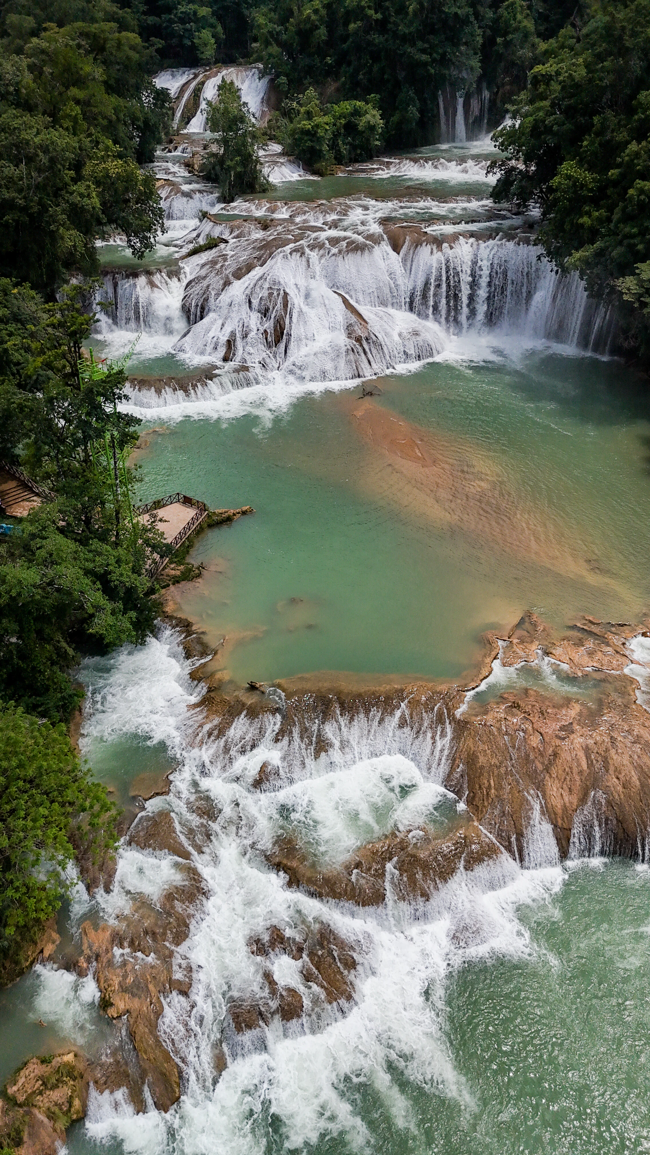 High-angle drone view of cascading waterfalls over rocky tiers into turquoise pools surrounded by lush green tropical forest.