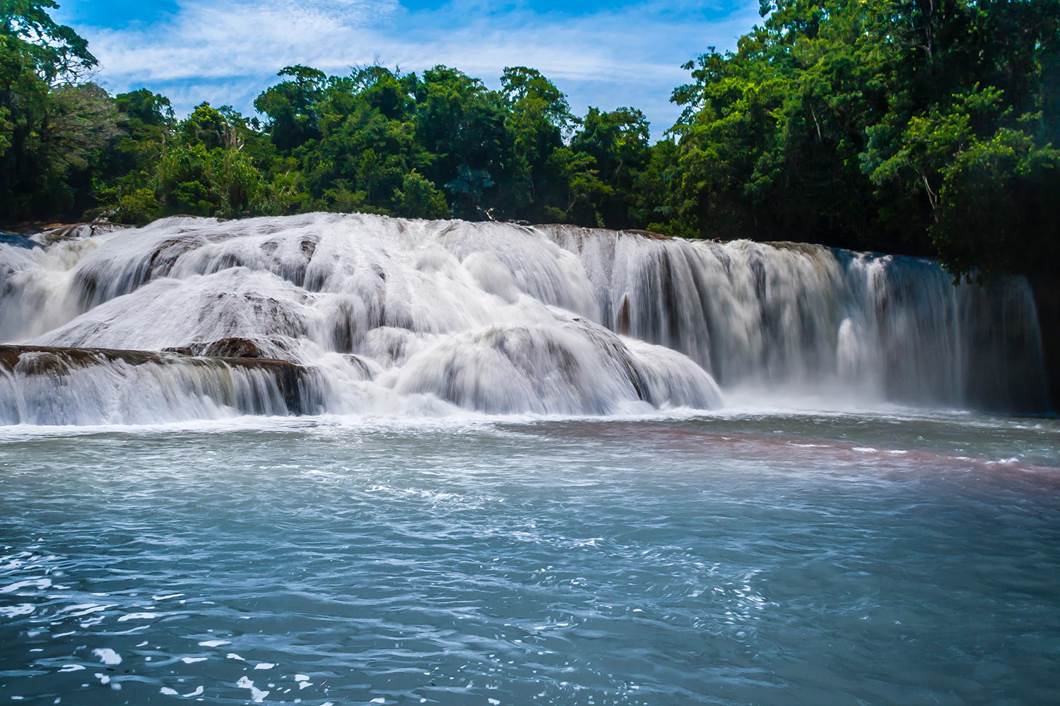 A wide, cascading waterfall flowing over limestone shelves into a turquoise pool, surrounded by dense green forest foliage under a partly cloudy blue sky.