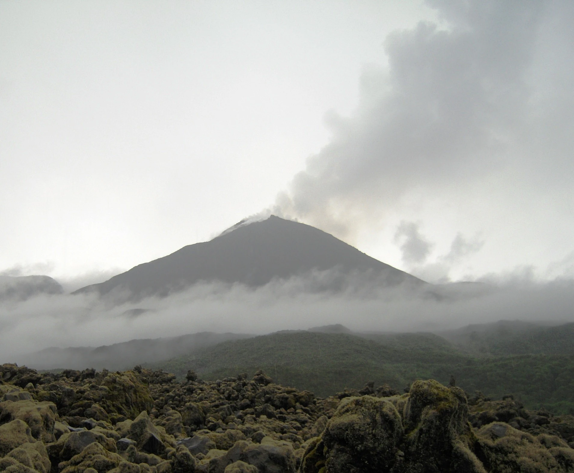 A wide shot of the conical El Reventador volcano under an overcast sky, with a plume of smoke and ash rising from the summit and low clouds surrounding the base, in front of a rocky volcanic foreground.