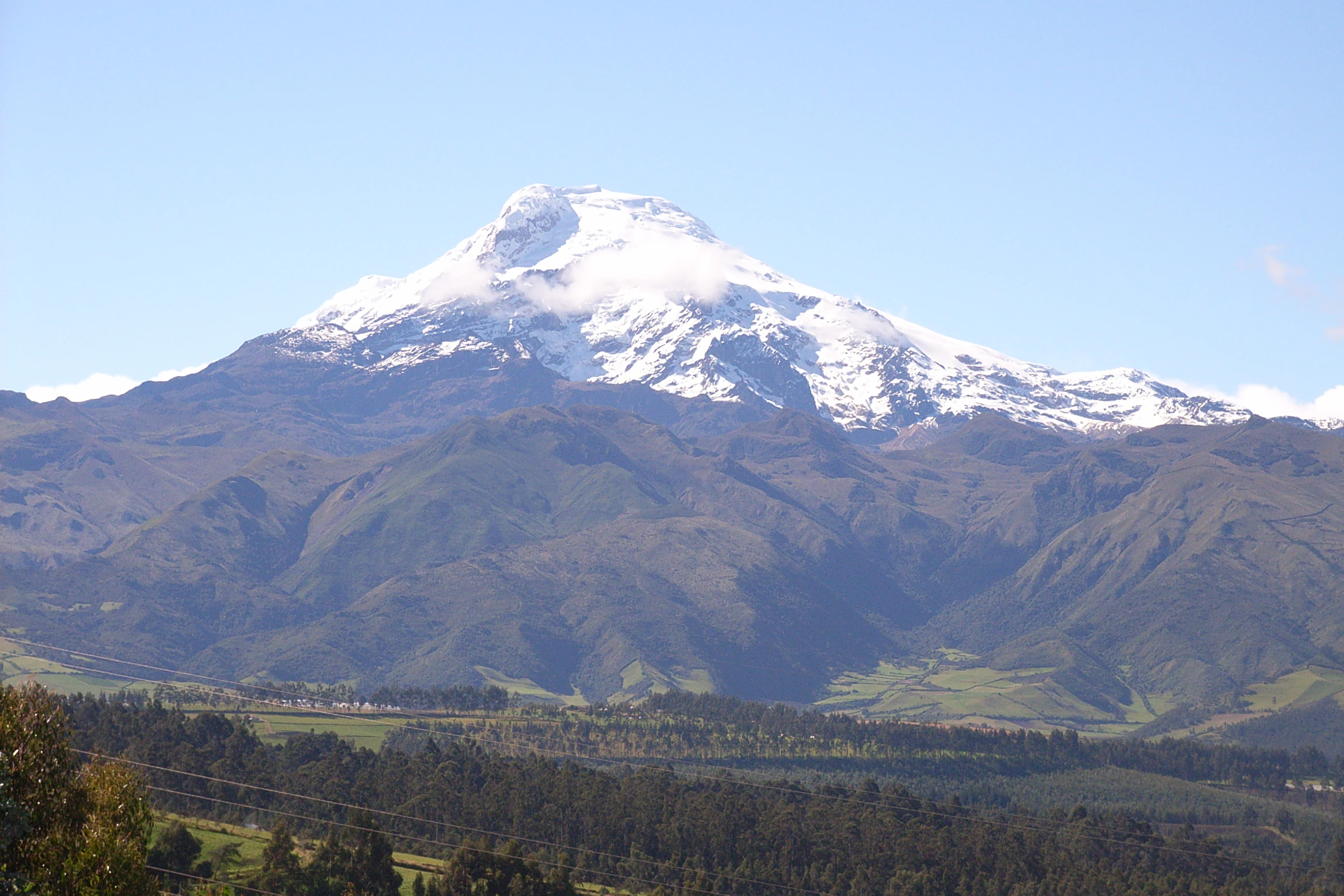 A wide shot of the snow-covered Cayambe volcano peak rising above green mountainous slopes and foothills.