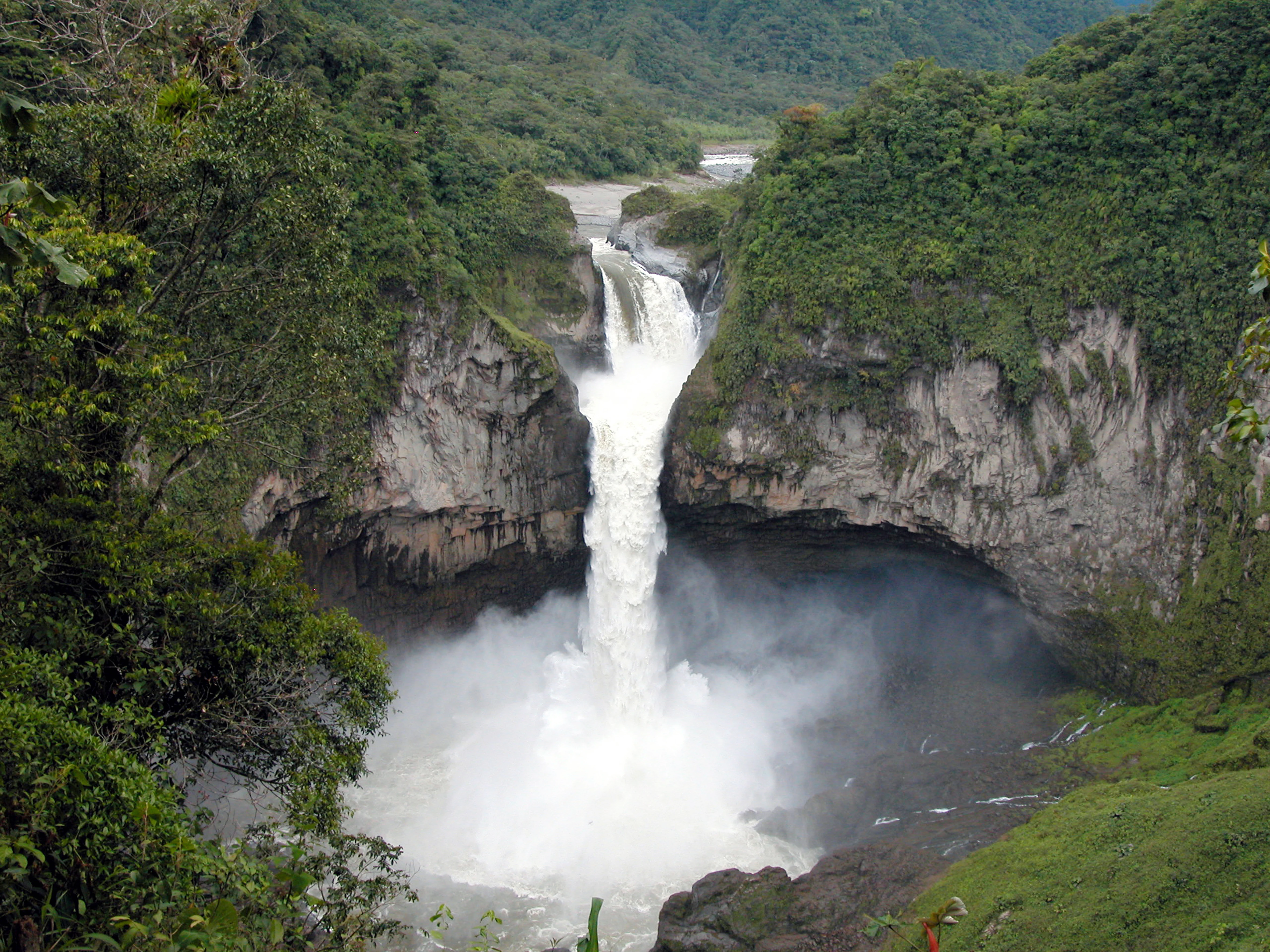 A high-angle view of a massive waterfall cascading over a vertical cliff into a misty pool, surrounded by dense green forest.