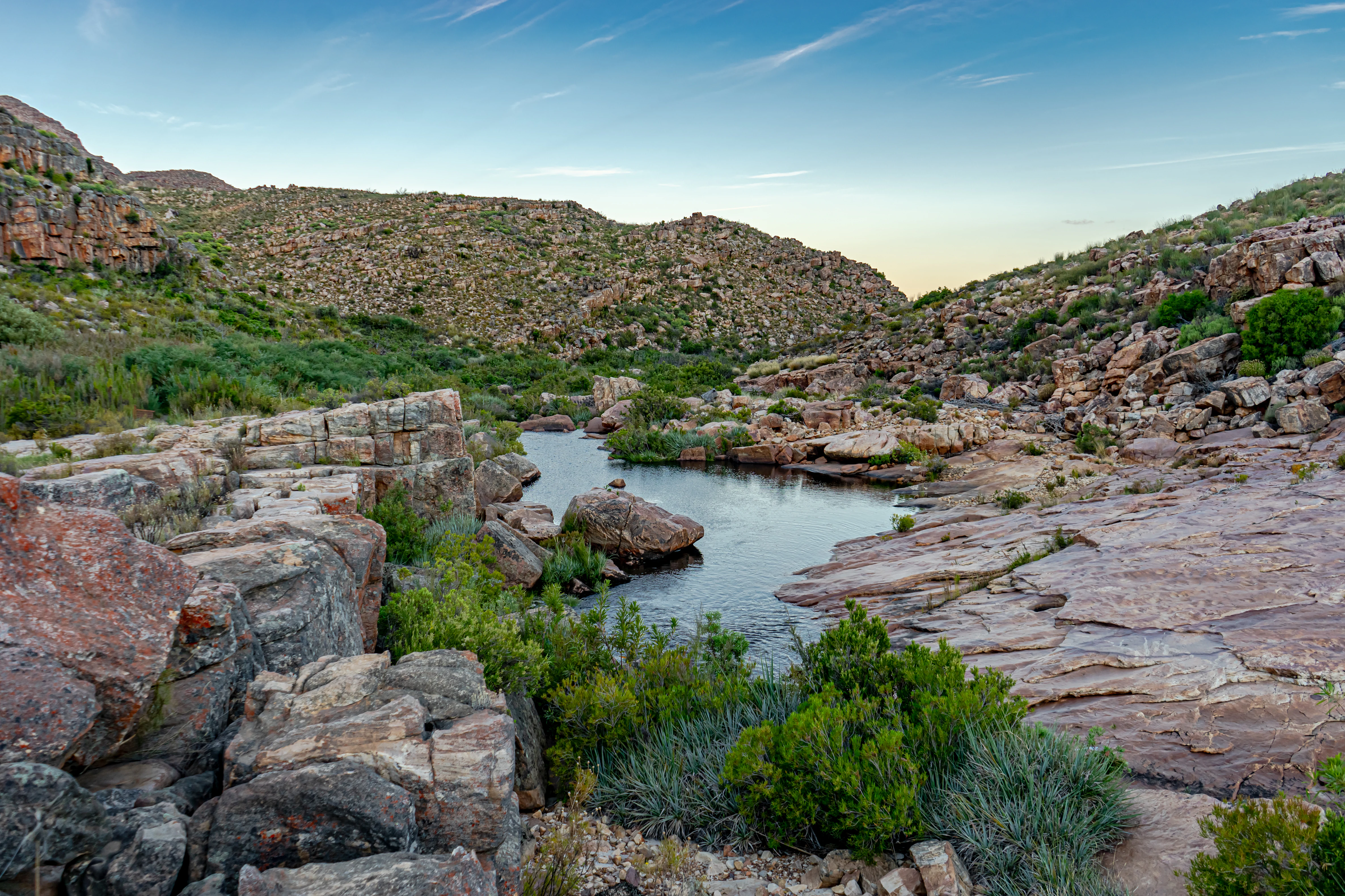 A small river pool sits amidst layered, weathered sandstone rocks and low-lying shrubs under a clear sky in a mountainous wilderness landscape.