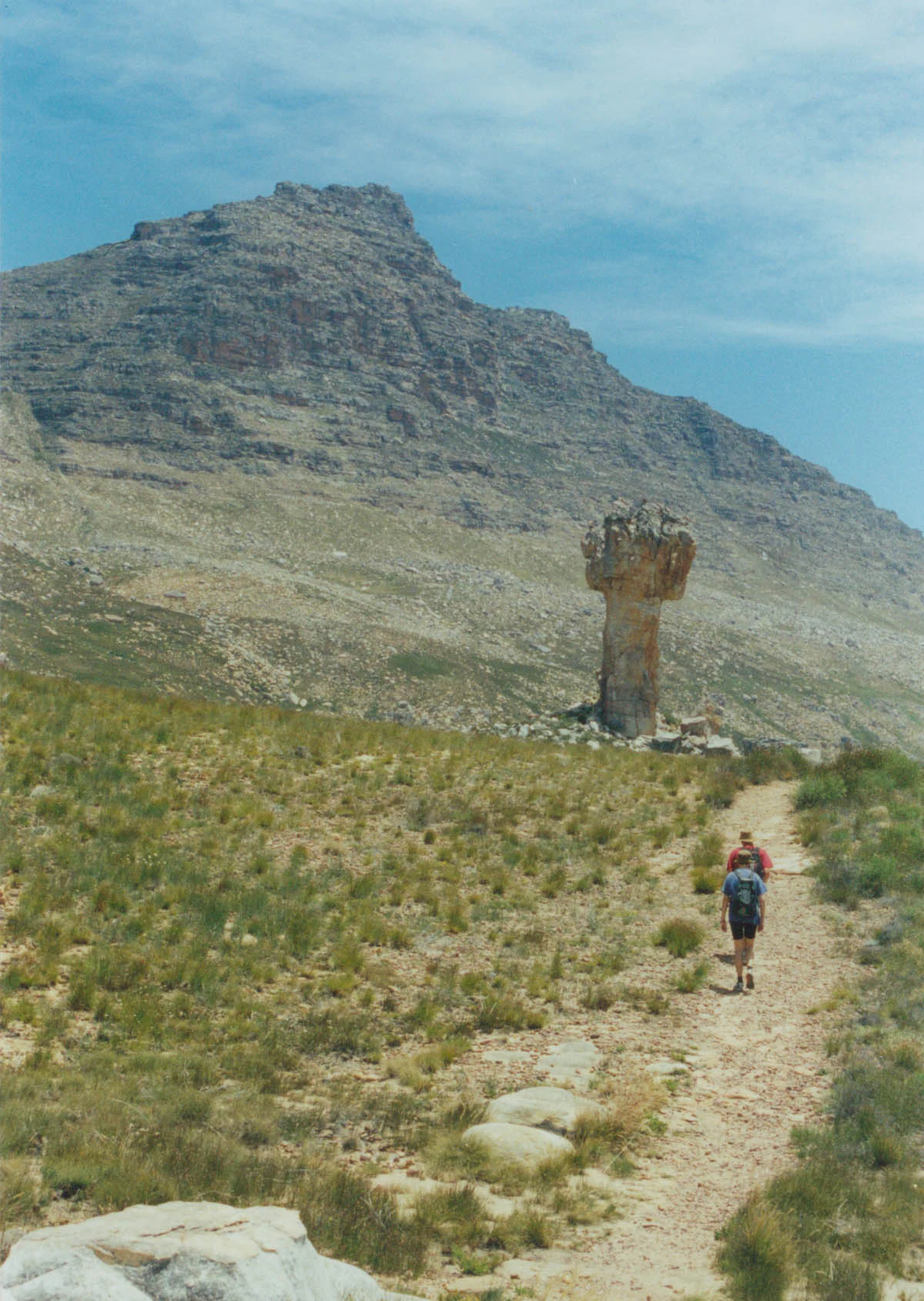 Two hikers walk on a dirt path through a grassy, arid landscape toward a large, cross-shaped sandstone rock formation, with a rugged mountain massif in the background.