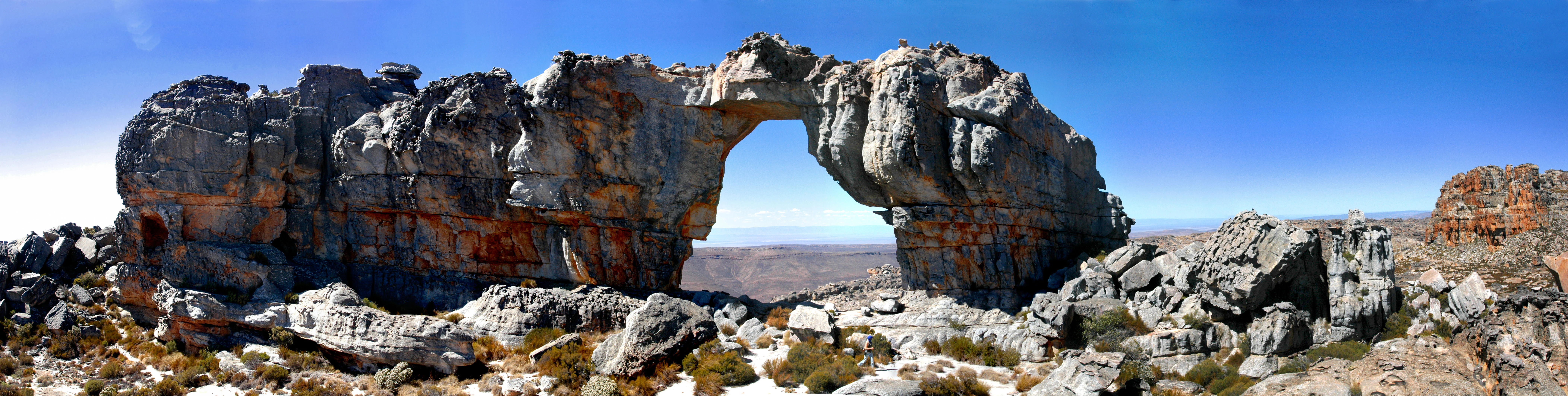 A wide panoramic view of the Wolfberg Arch, a large natural sandstone rock arch formation set in a rugged mountainous desert landscape under a bright blue sky.