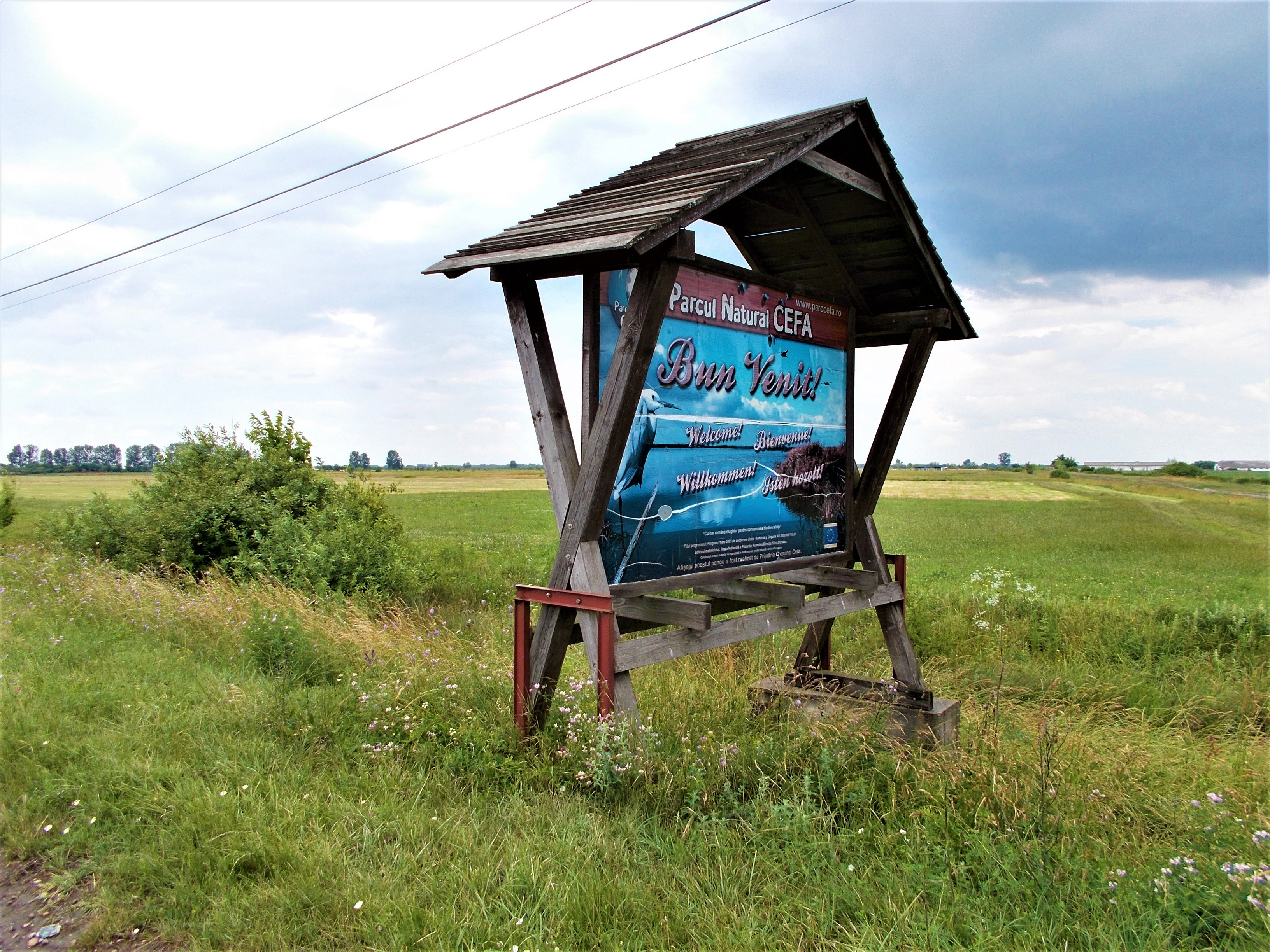 Wooden entrance sign structure with "Parcul Natural Cefa" text, situated in a grassy field with open landscape in background