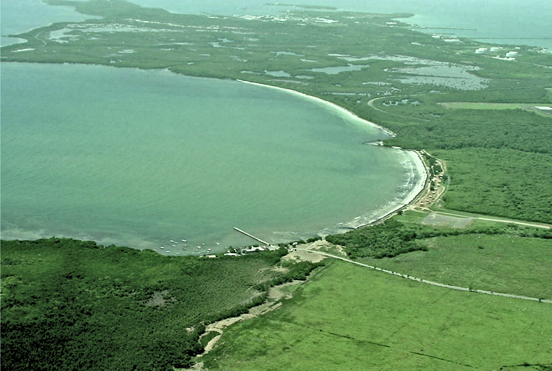 Fotografía aérea de un paisaje costero que muestra una gran laguna curva de aguas color verde turquesa, una costa de arena, un denso bosque de manglares y una carretera costera que recorre la orilla.