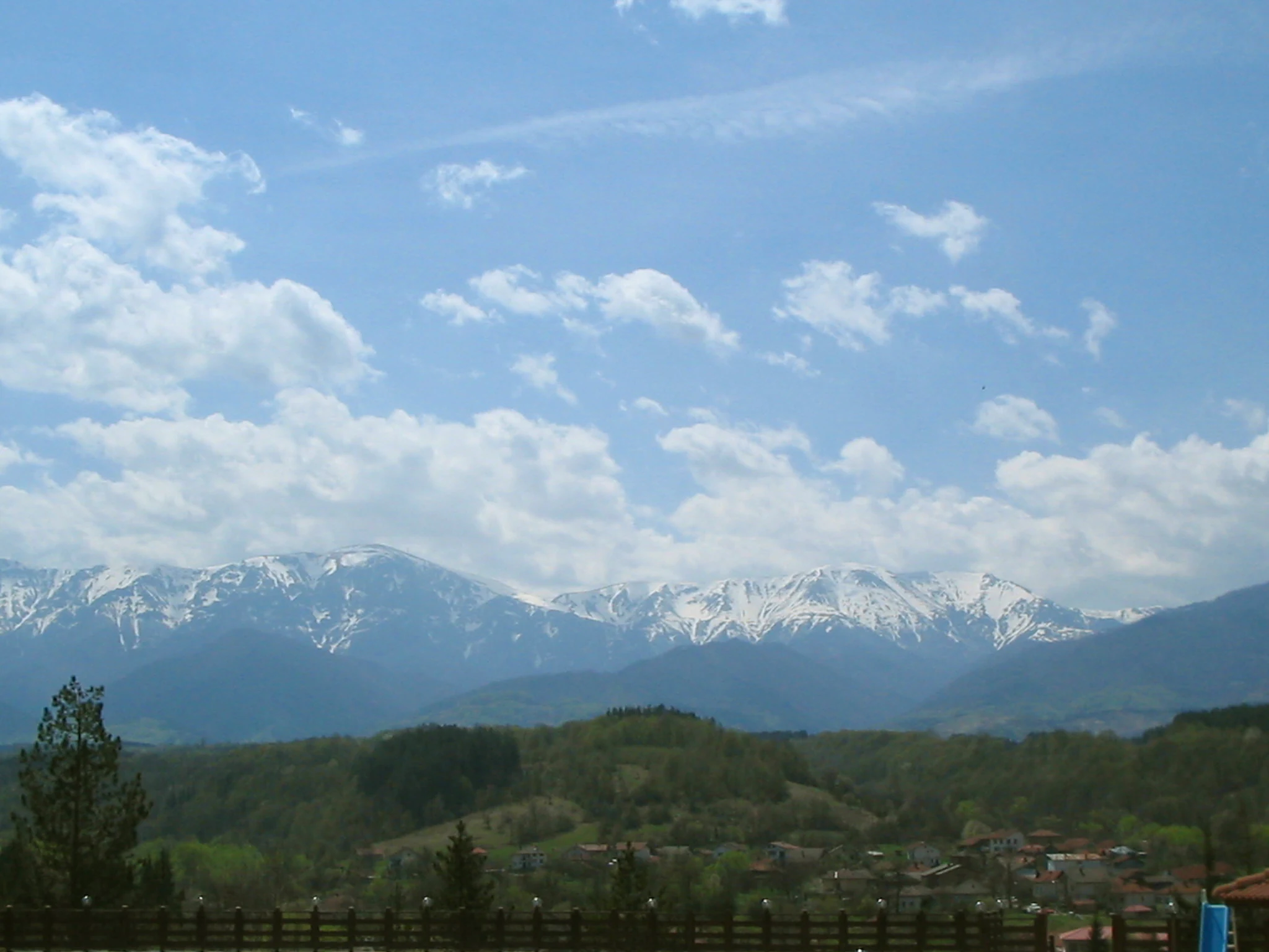 Snow-covered mountain peaks under a partly cloudy sky, with green hills and a village in the foreground