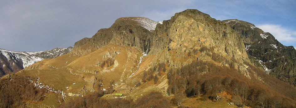 Mountainous landscape with rocky peaks, patches of snow, and a waterfall in the distance under a partly cloudy sky