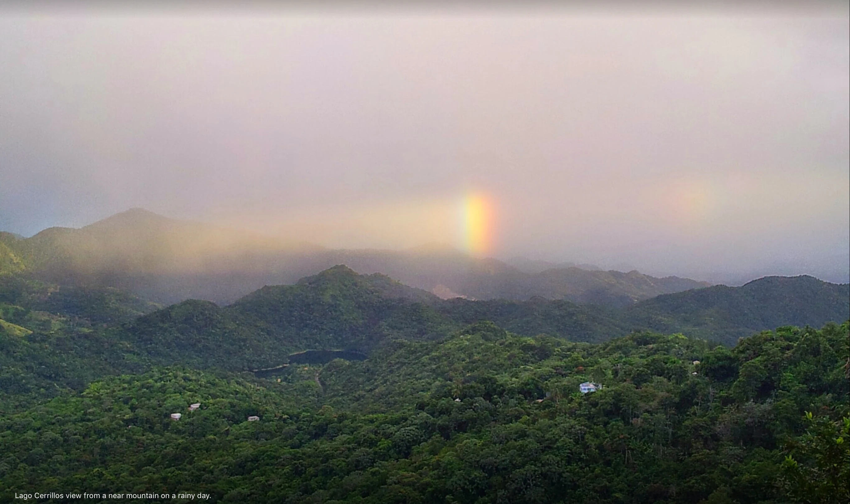 A wide, slightly hazy, eye-level landscape view of rolling, green, forested mountains under a cloudy, overcast sky with a faint rainbow segment hanging above the ridges.