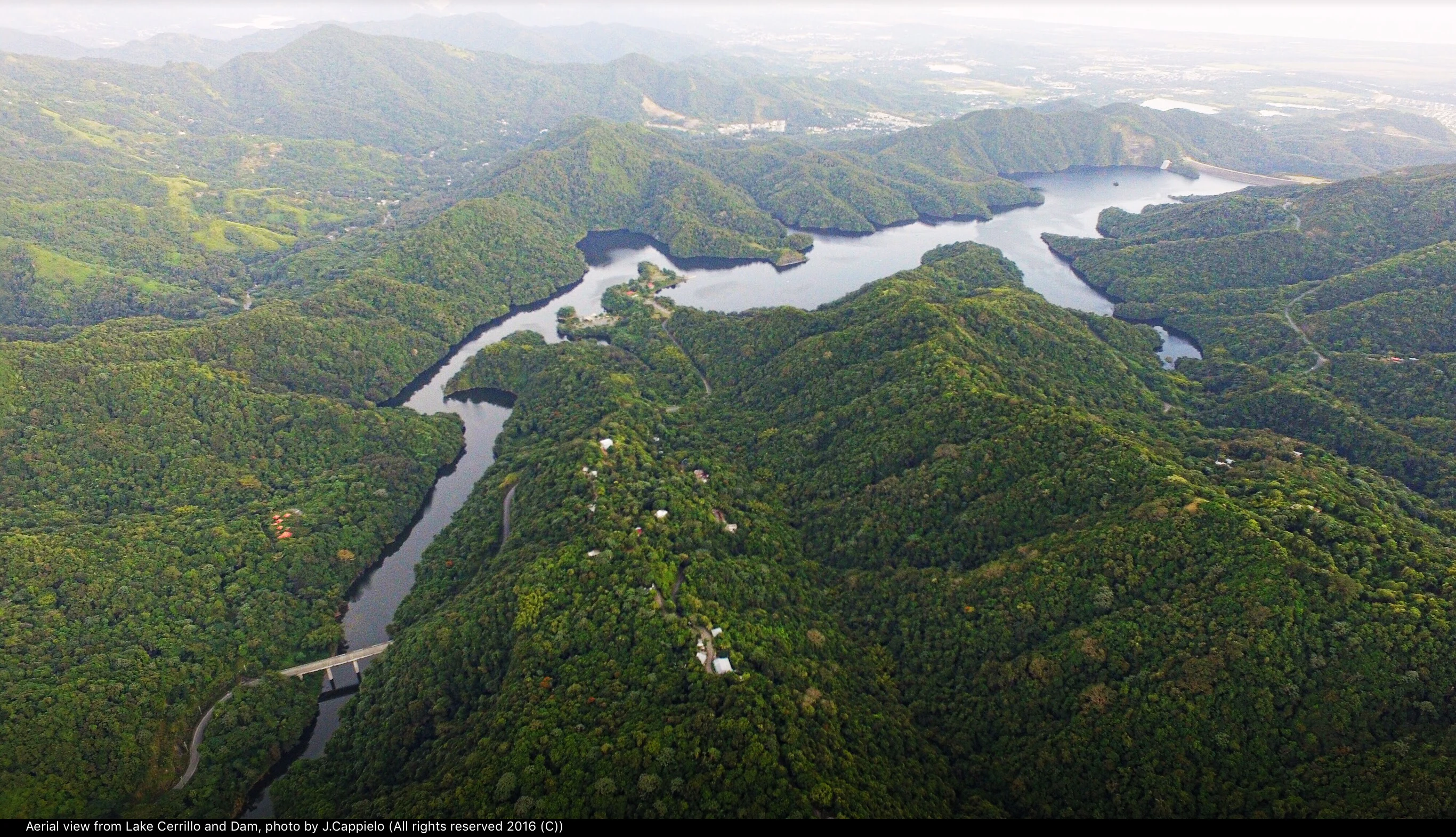 Aerial view of a winding lake surrounded by dense, green forest-covered mountains, with a dam structure visible in the lower foreground.