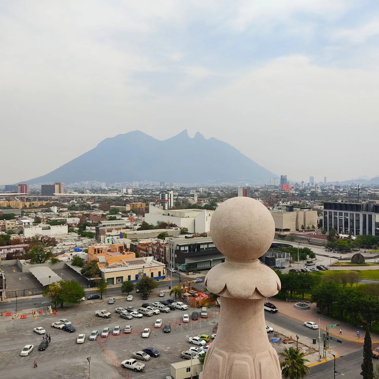 A wide angle shot of the distinctive saddle shaped mountain Cerro de la Silla in the distance, framed by a concrete architectural baluster in the foreground and the sprawling urban environment of Monterrey below.