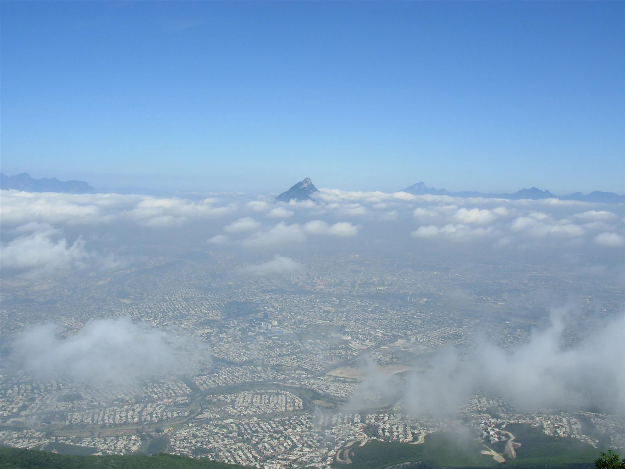 A high angle view looking down over a large city spread out beneath a layer of clouds, with a distant mountain peak visible above the cloud line.