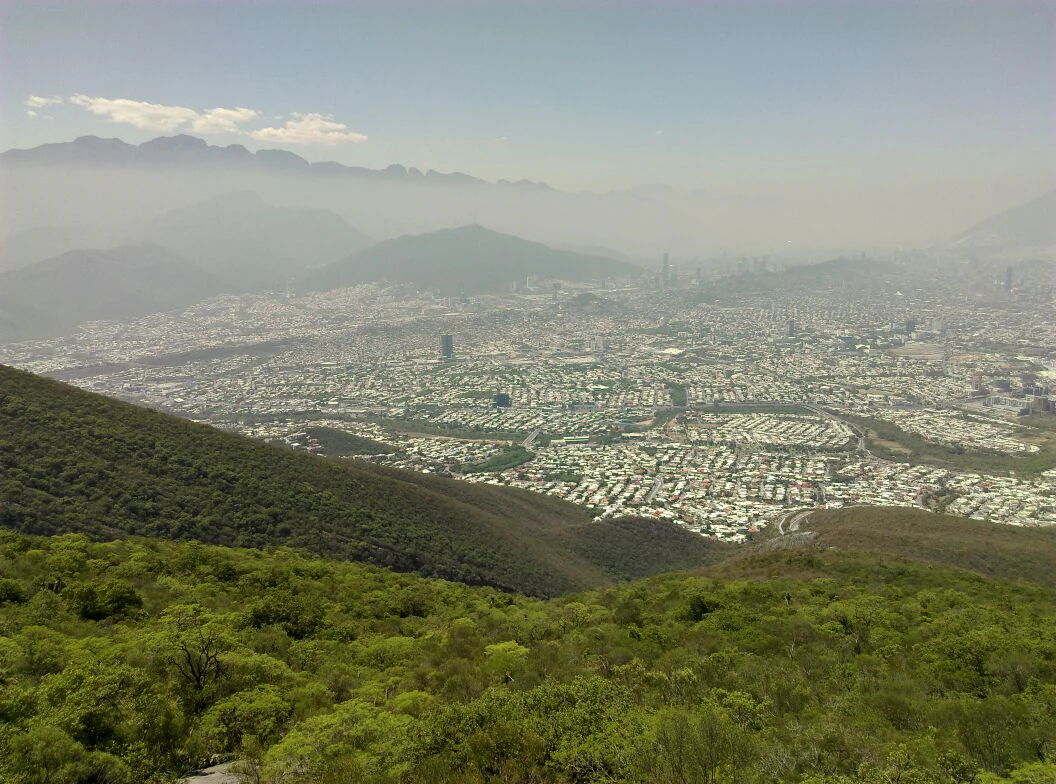A wide angle view from the mountain slopes looking down at the sprawling city of Monterrey, Mexico, partially obscured by hazy air under a light sky.