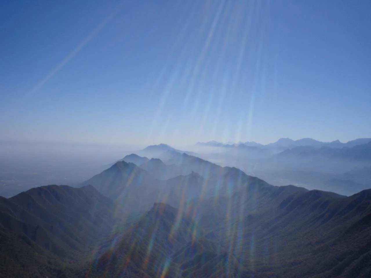A wide, hazy landscape shot looking out over a series of mountain ridges under a bright, sunny sky with lens flare.