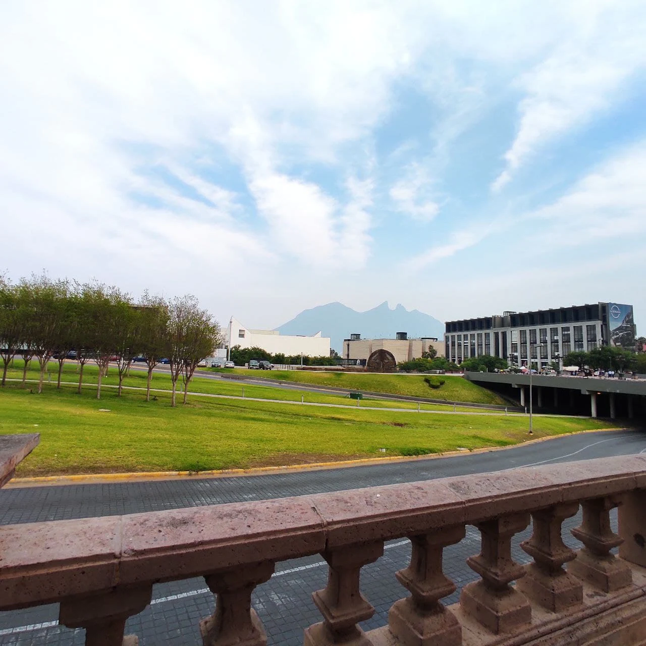 A view across a grassy park and road towards the prominent ridgeline of Cerro de la Silla in the distance, framed by a stone balustrade in the foreground.