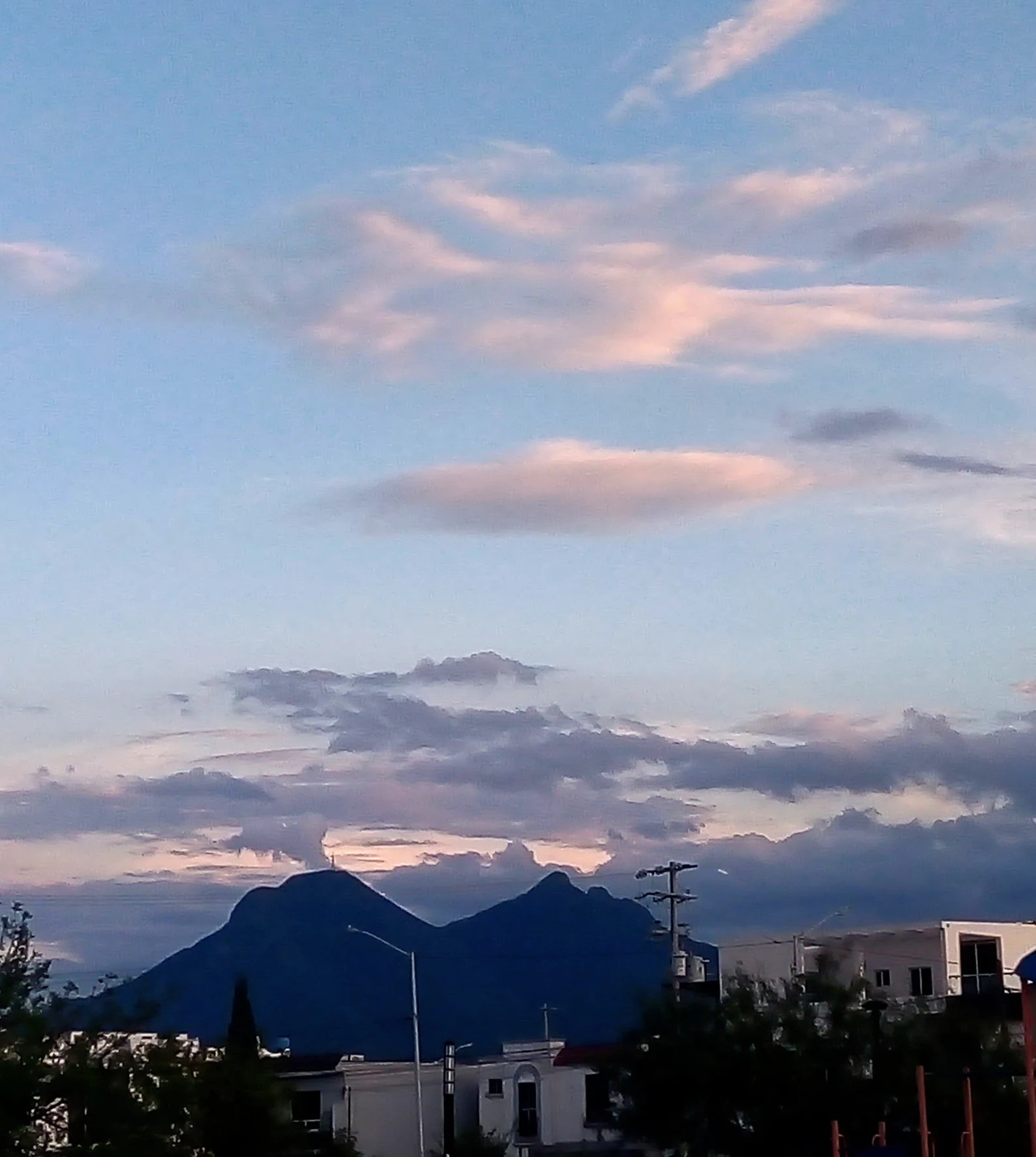 A view of the distinctive saddle-shaped peaks of Cerro de la Silla mountain against a twilight sky with scattered lenticular and cumulus clouds, framed by suburban buildings and trees in the foreground.