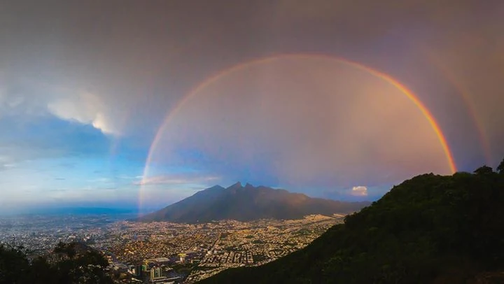 A wide view of the Cerro de la Silla mountain rising above the city of Monterrey with a bright double rainbow spanning the sky.