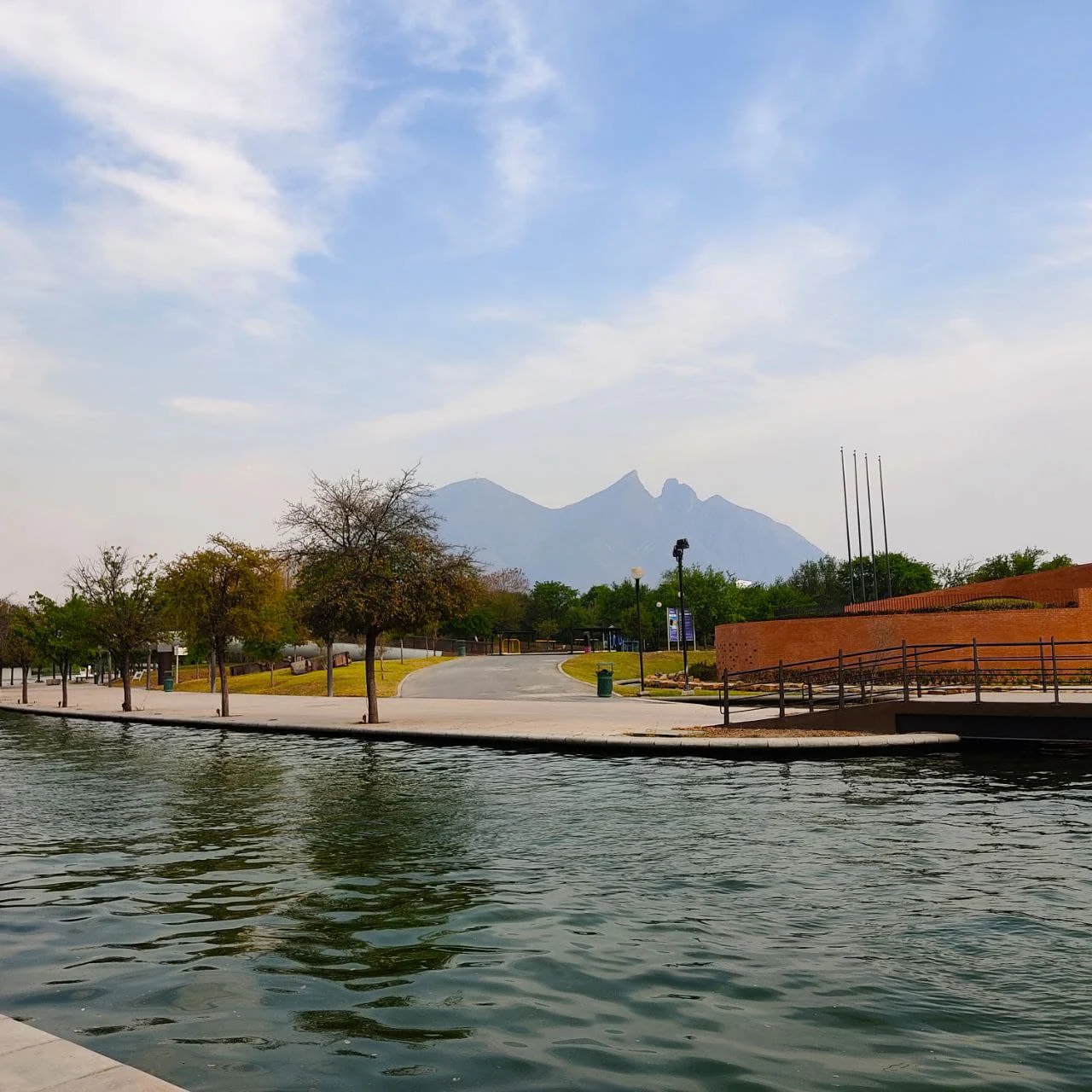 A wide angle view of the distinctive Cerro de la Silla mountain peaks in the background, overlooking a canal with a concrete pathway, trees, and brick structures in the foreground.