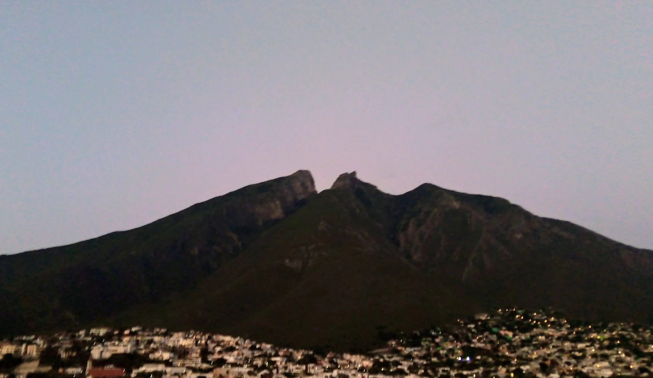A wide shot of Cerro de la Silla mountain under a soft sky, with urban development visible in the foreground.