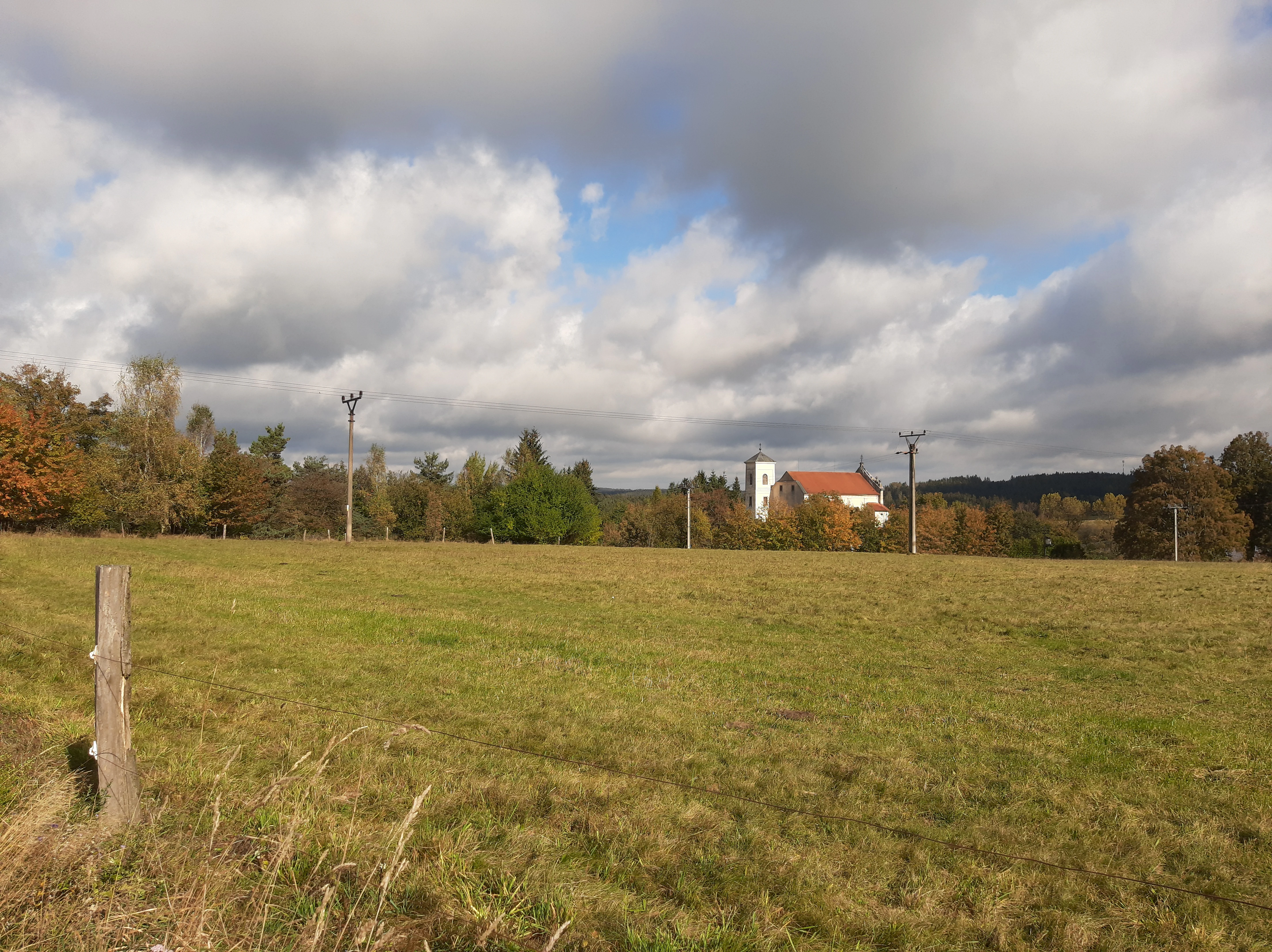A wide view of a grassy field with a monastery building nestled among trees in the distance under a cloudy sky.